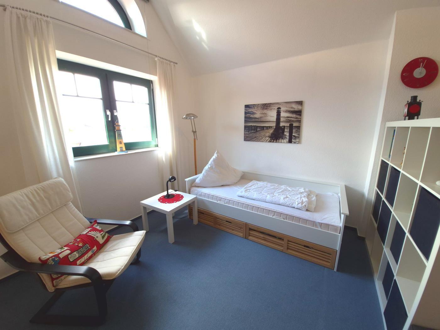 Schlafzimmer mit Bett, Sessel und Fenster. Wandbild und Regal sind sichtbar.