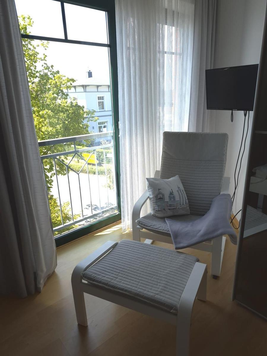 Ein Raum mit Sessel, Fußstütze und TV. Durchgehendes Fenster mit Blick auf Balkon und Grün.