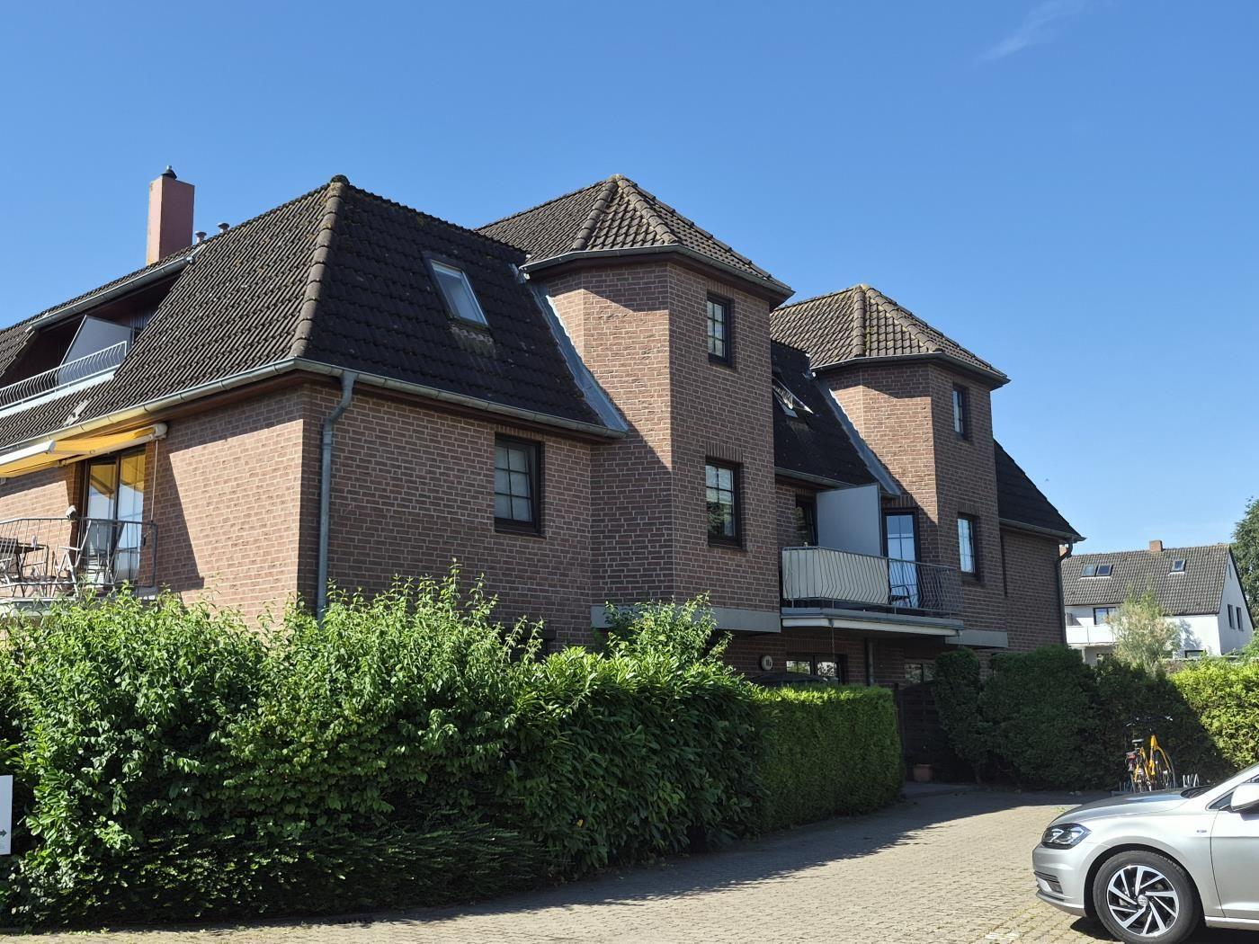 Zweistöckiges Backsteinhaus mit Balkonen und grüner Hecke. Parkplatz mit Auto.