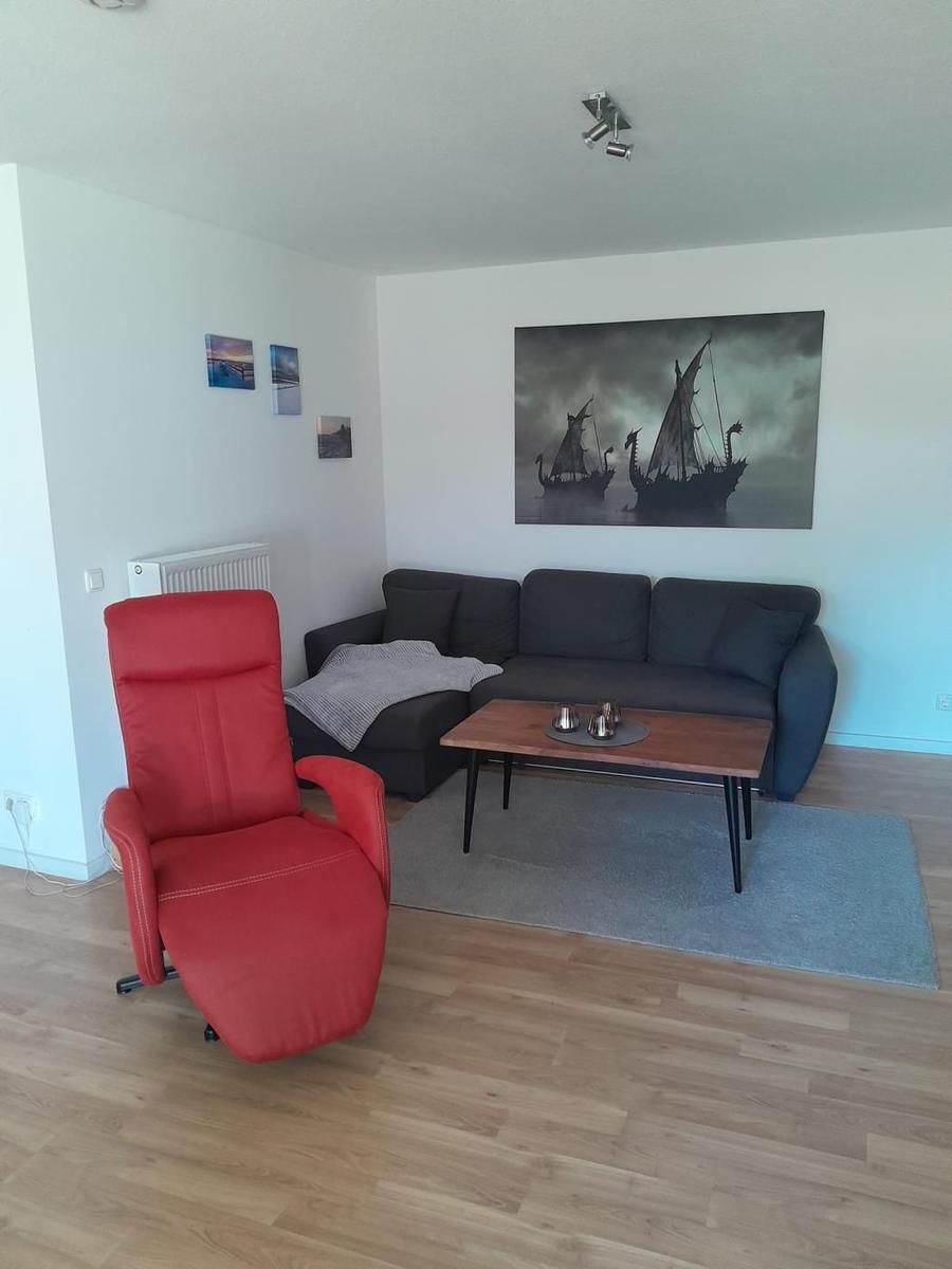 Ein Wohnzimmer mit grauem Sofa, rotem Sessel und Holzboden. An der Wand hängt ein großes Bild von Schiffen.
