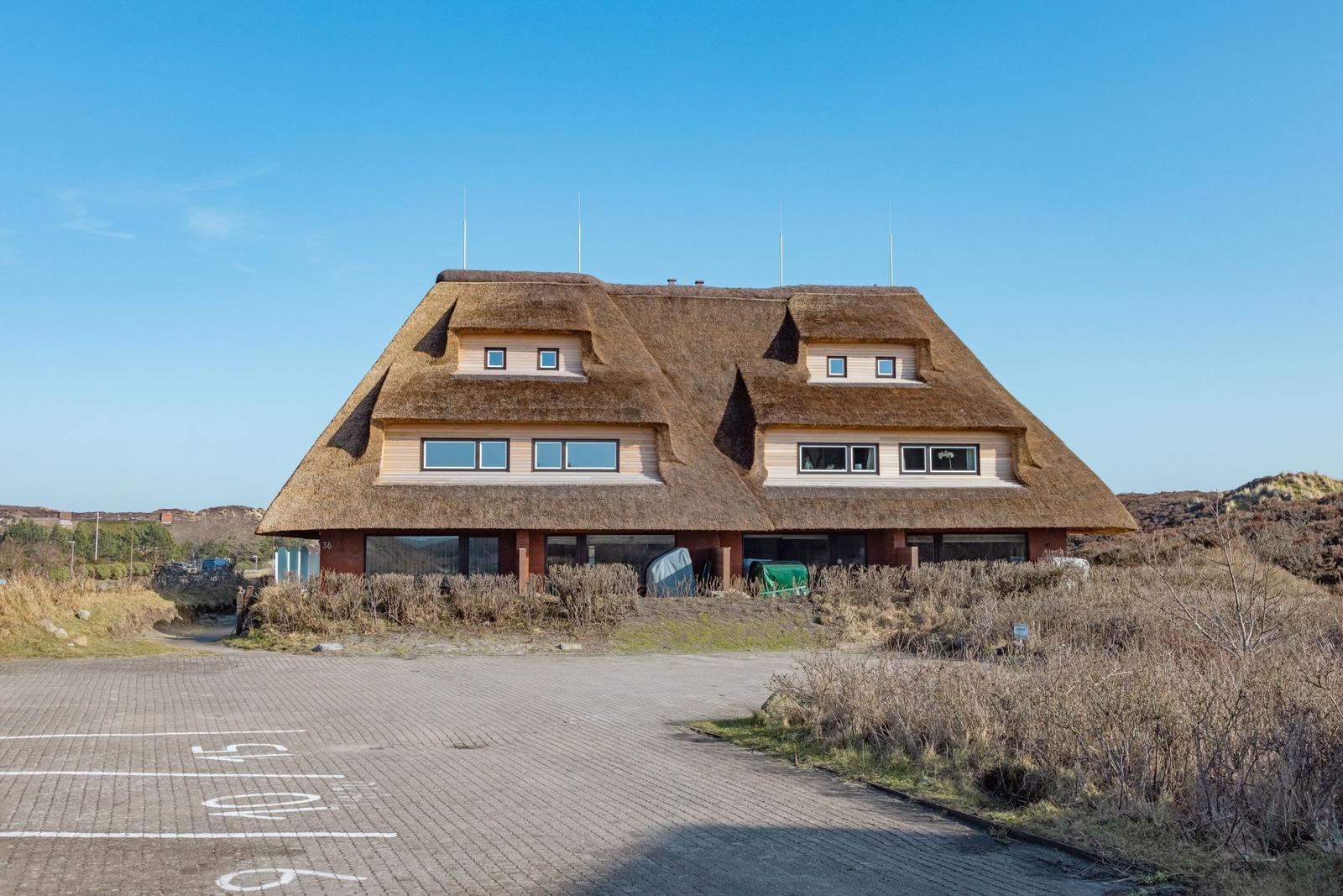 Haus mit strohgedecktem Dach und großen Fenstern in ländlicher Umgebung.