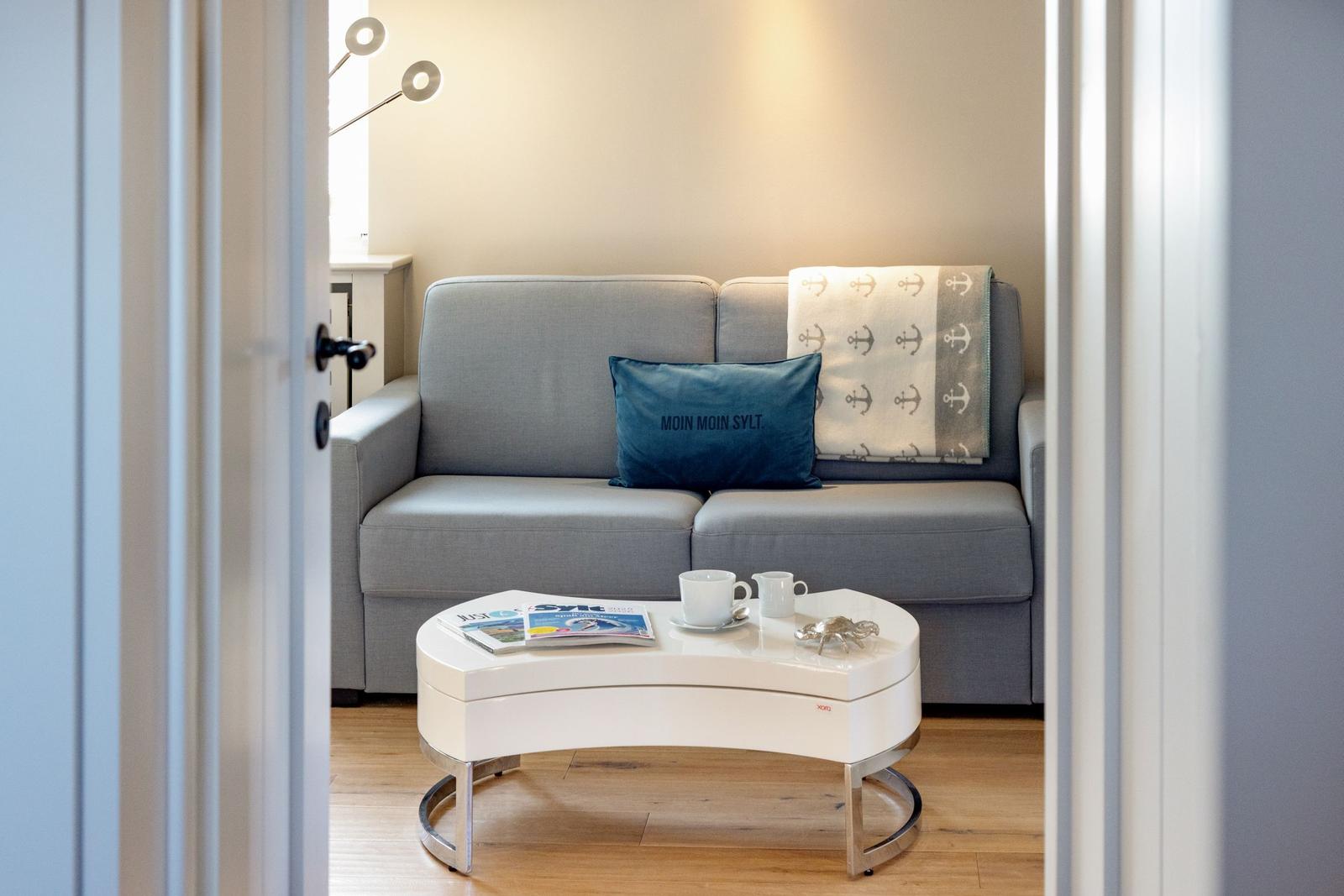 Ein grauer Sofa mit Kissen und Decke steht vor einem weißen Tisch mit Tassen und Zeitschriften.
