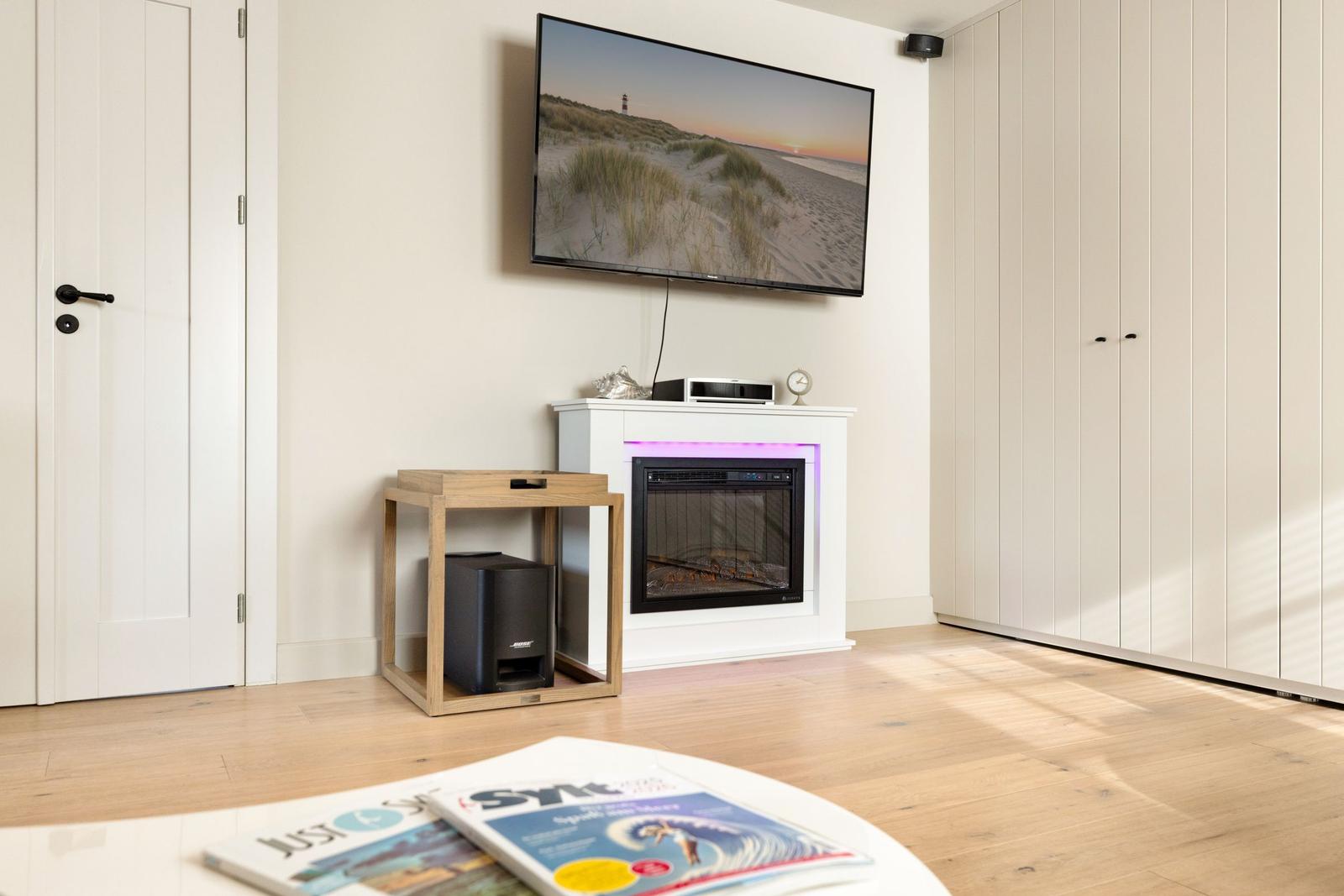 Wohnzimmer mit Fernseher, Kamin und Holzboden. Links Tür, rechts Schrank.