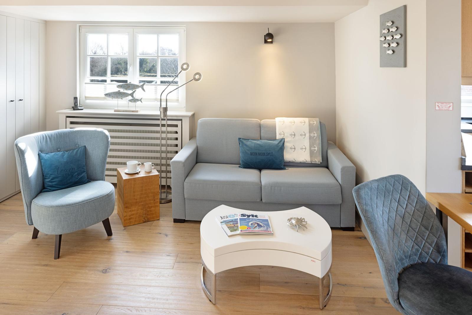 Wohnzimmer mit Sofa, Sessel und Tisch. Fenster, Wandlampe und Boden sind sichtbar.