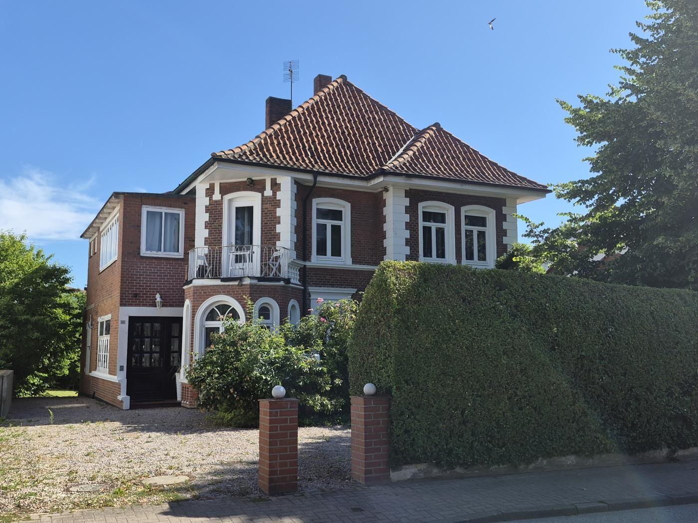 Zweistöckiges Haus mit rotem Ziegeldach und weißen Fensterrahmen. Vorgarten mit Hecke und Steinweg.