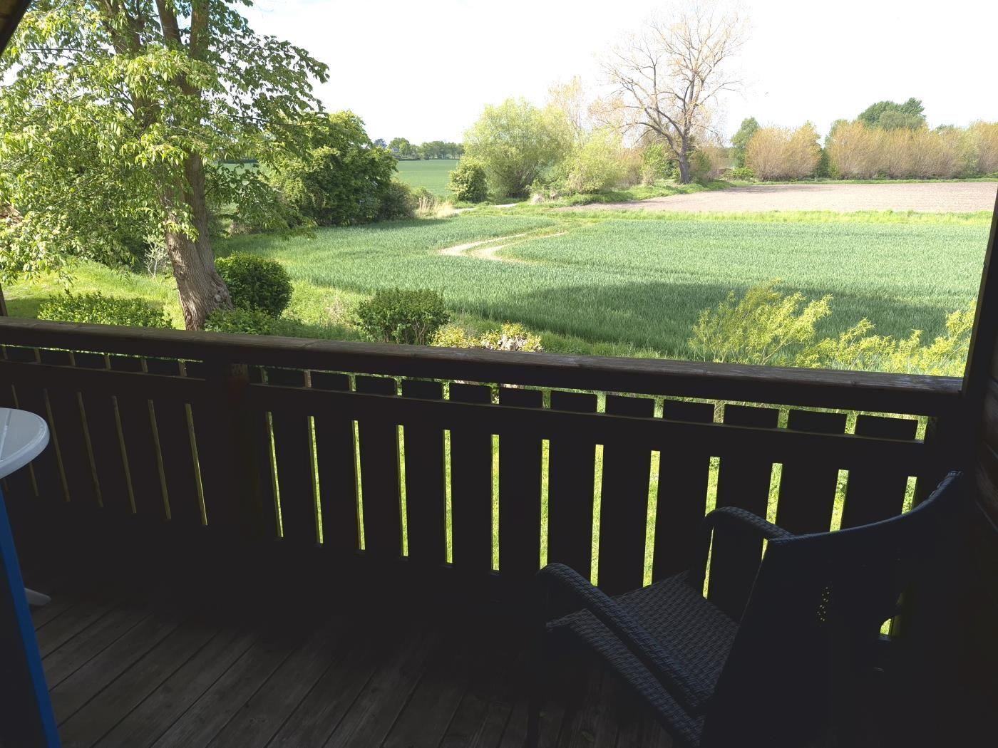Terrasse mit Blick auf grüne Wiesen und Bäume. Holzgeländer und Stuhl sind sichtbar.