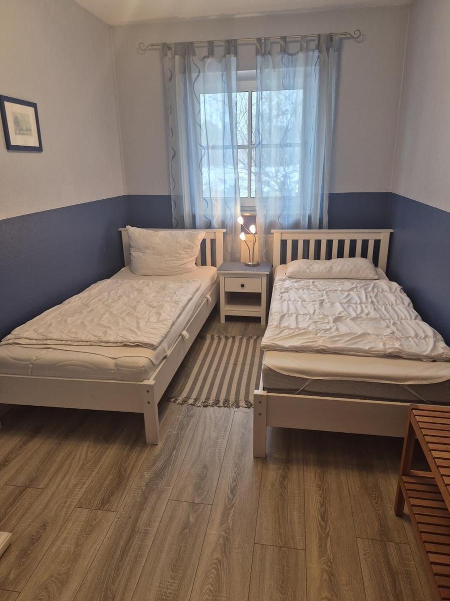 Zwei Einzelbetten, Nachttisch mit Lampe, Fenster mit Vorhängen, Holzboden und Wandgestaltung.