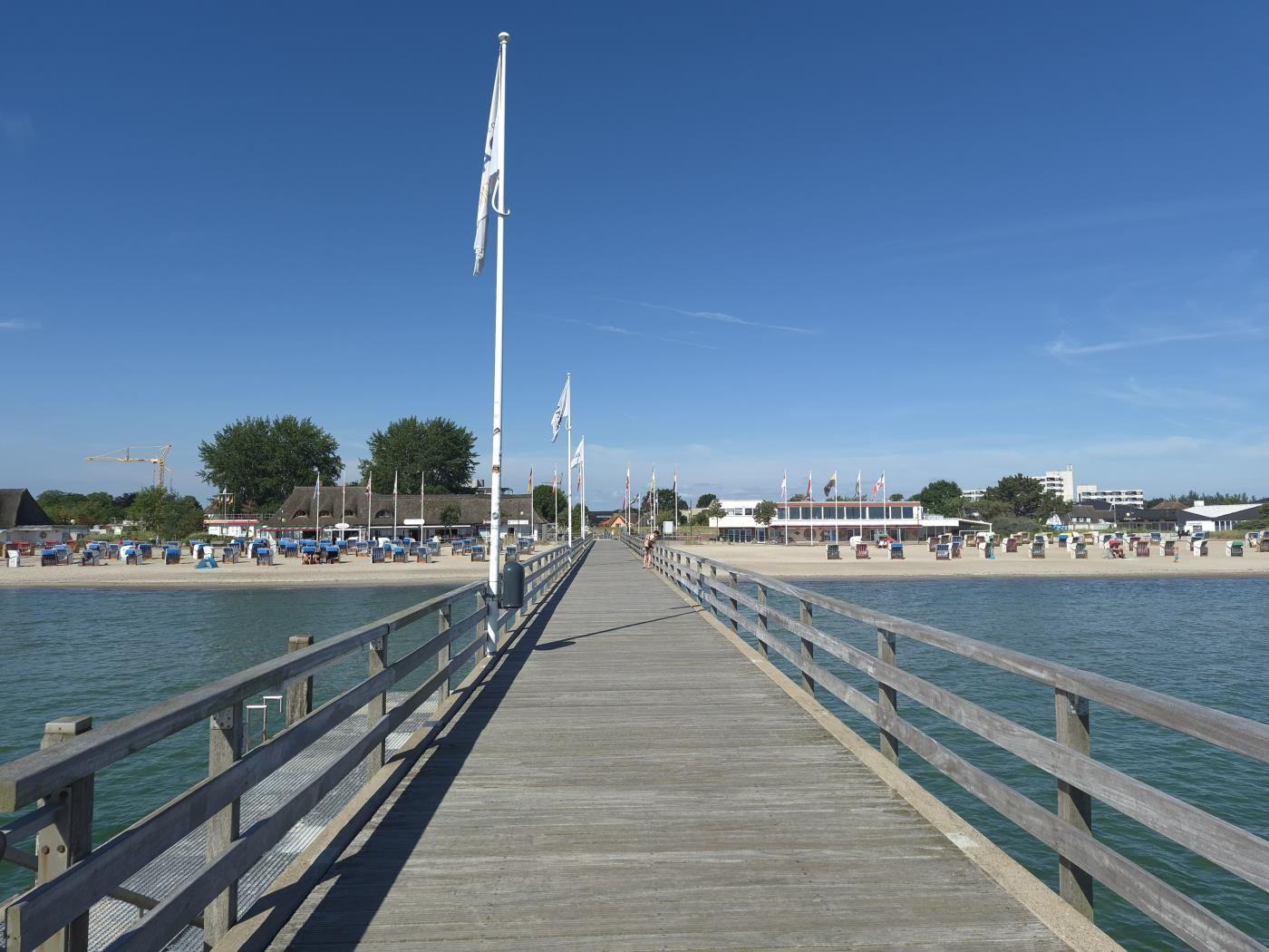 Holzbrücke führt zum Strand mit Strandkörben und Gebäuden im Hintergrund.