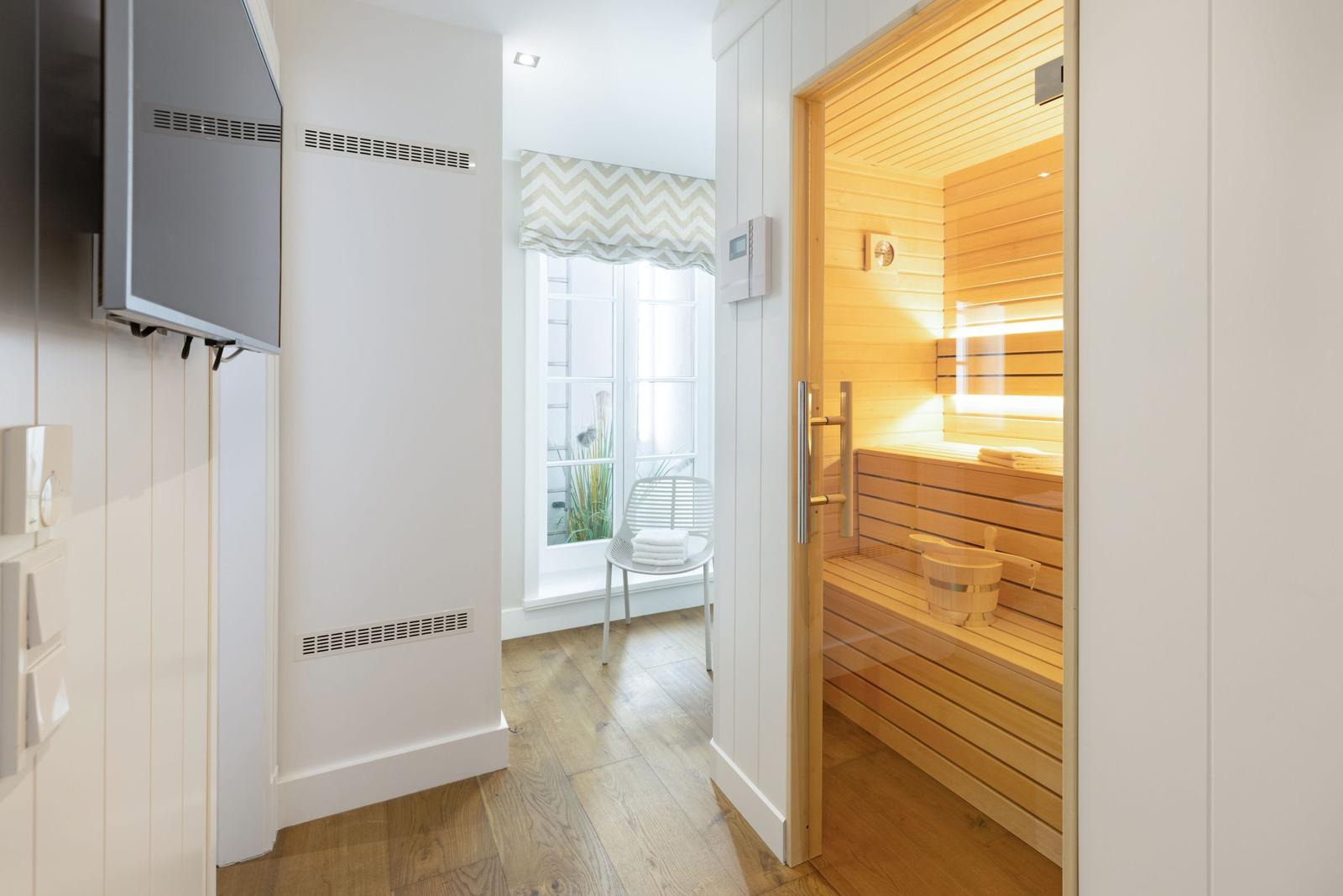 Sauna mit Holzbank und Eimer. Türöffnung zeigt Raum mit Fenster und Stuhl.
