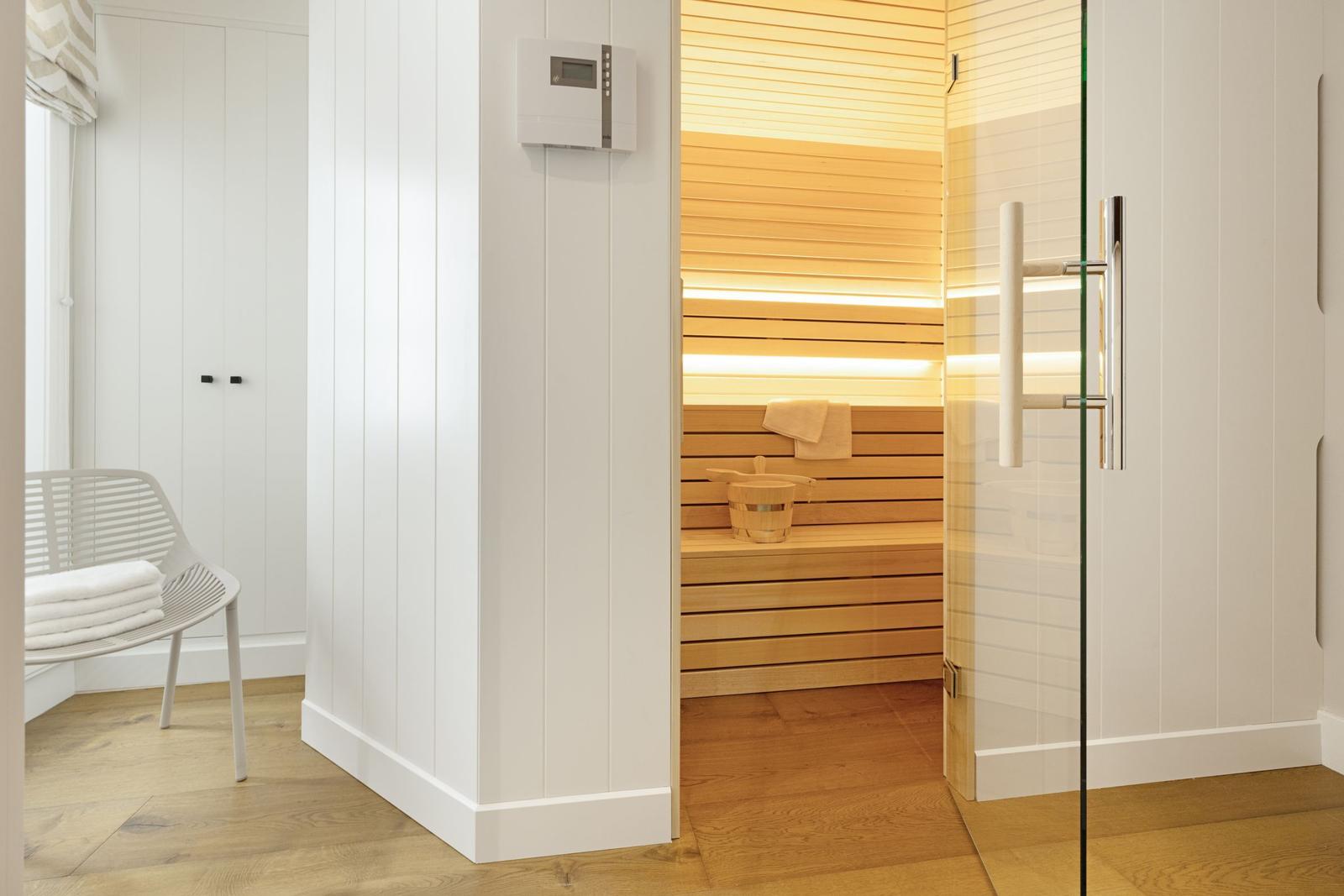 Sauna mit Holzbank und Glaswand. Weiße Wände und Holzboden. Dampfheizung an der Wand.
