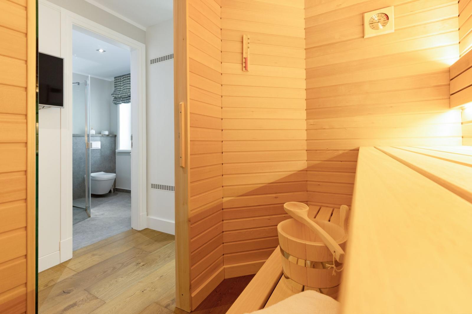 Sauna mit Holzbank und Eimer. Im Hintergrund ist ein Badezimmer mit Toilette und Dusche.