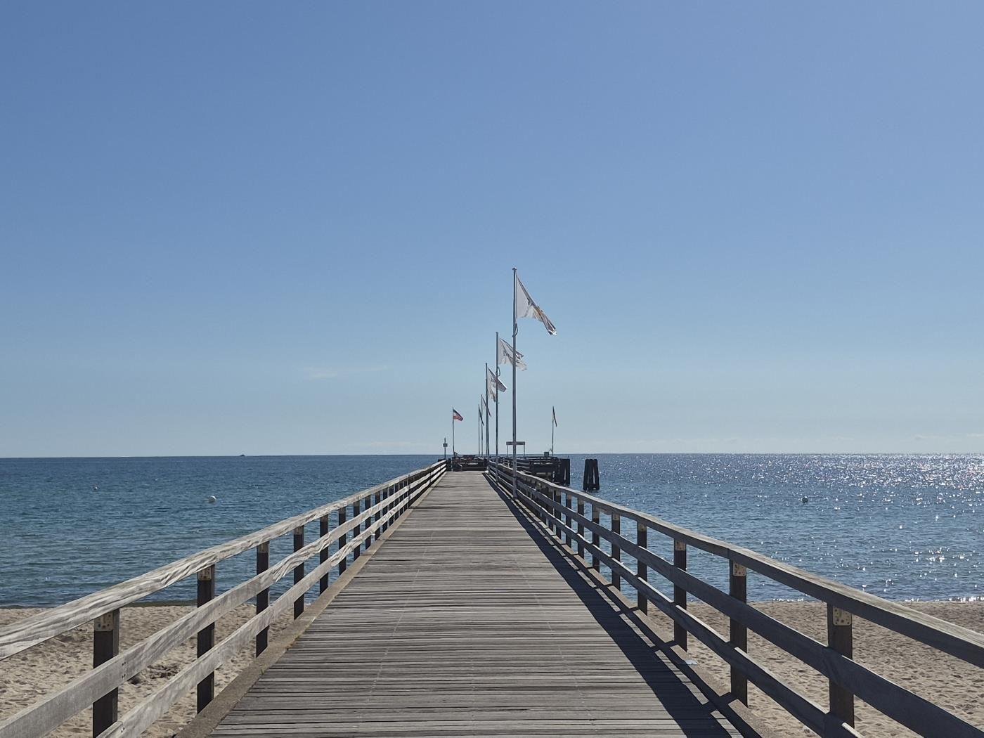 Holzsteg führt über Sandstrand zum Meer. Fahnen wehen im Wind.