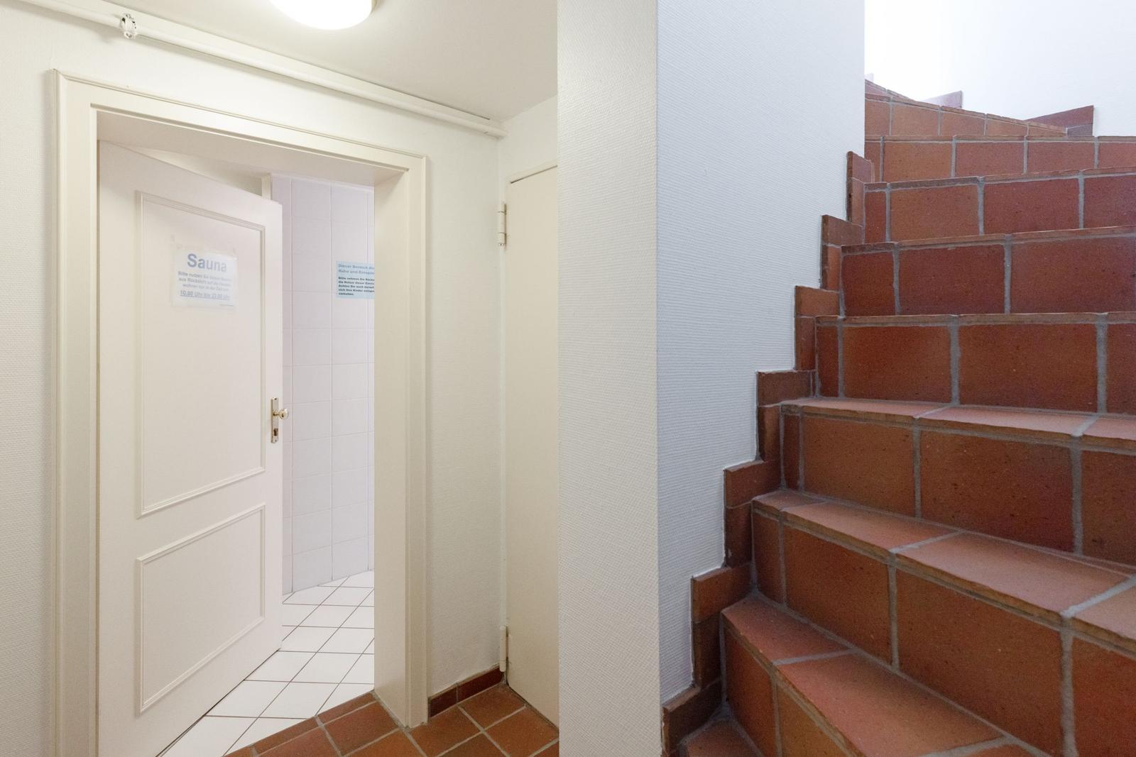 Sauna mit Tür und Fliesenboden. Rechts führt eine Backstein-Treppe nach oben.
