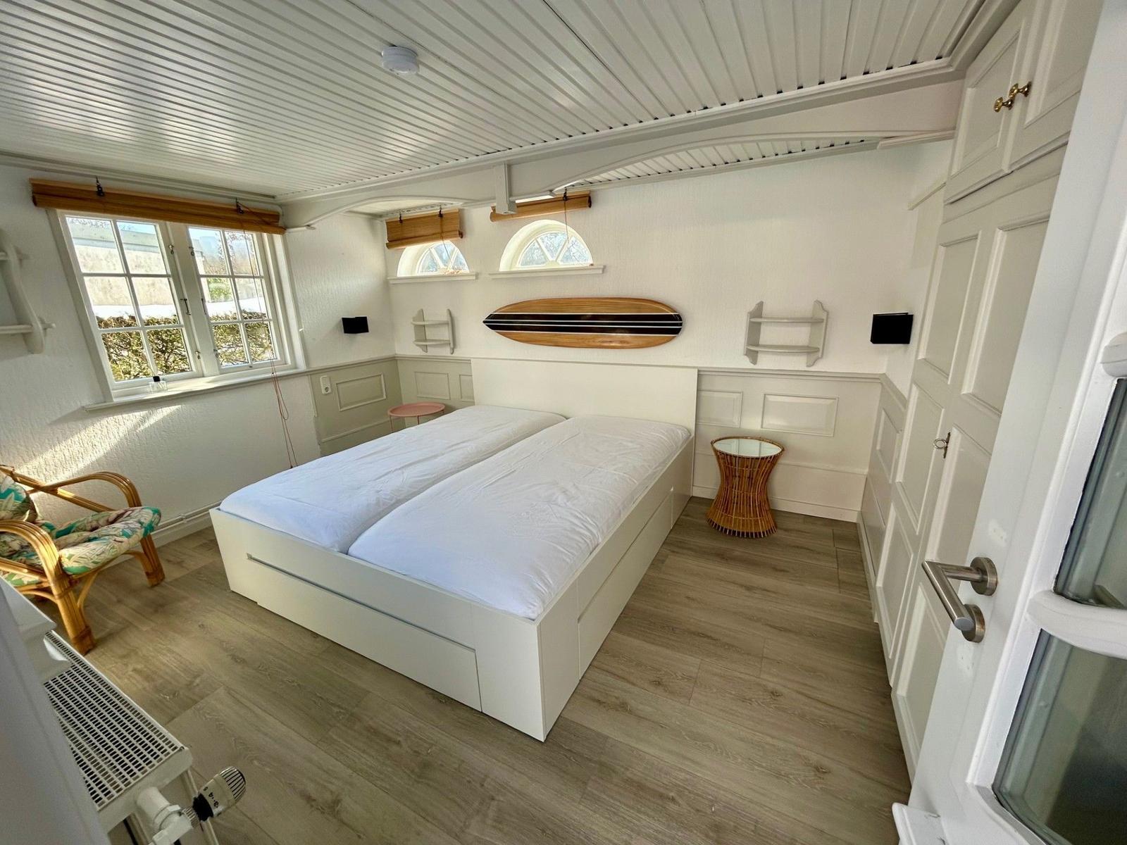 Schlafzimmer mit Doppelbett, Fenstern und Surfbrett an der Wand.