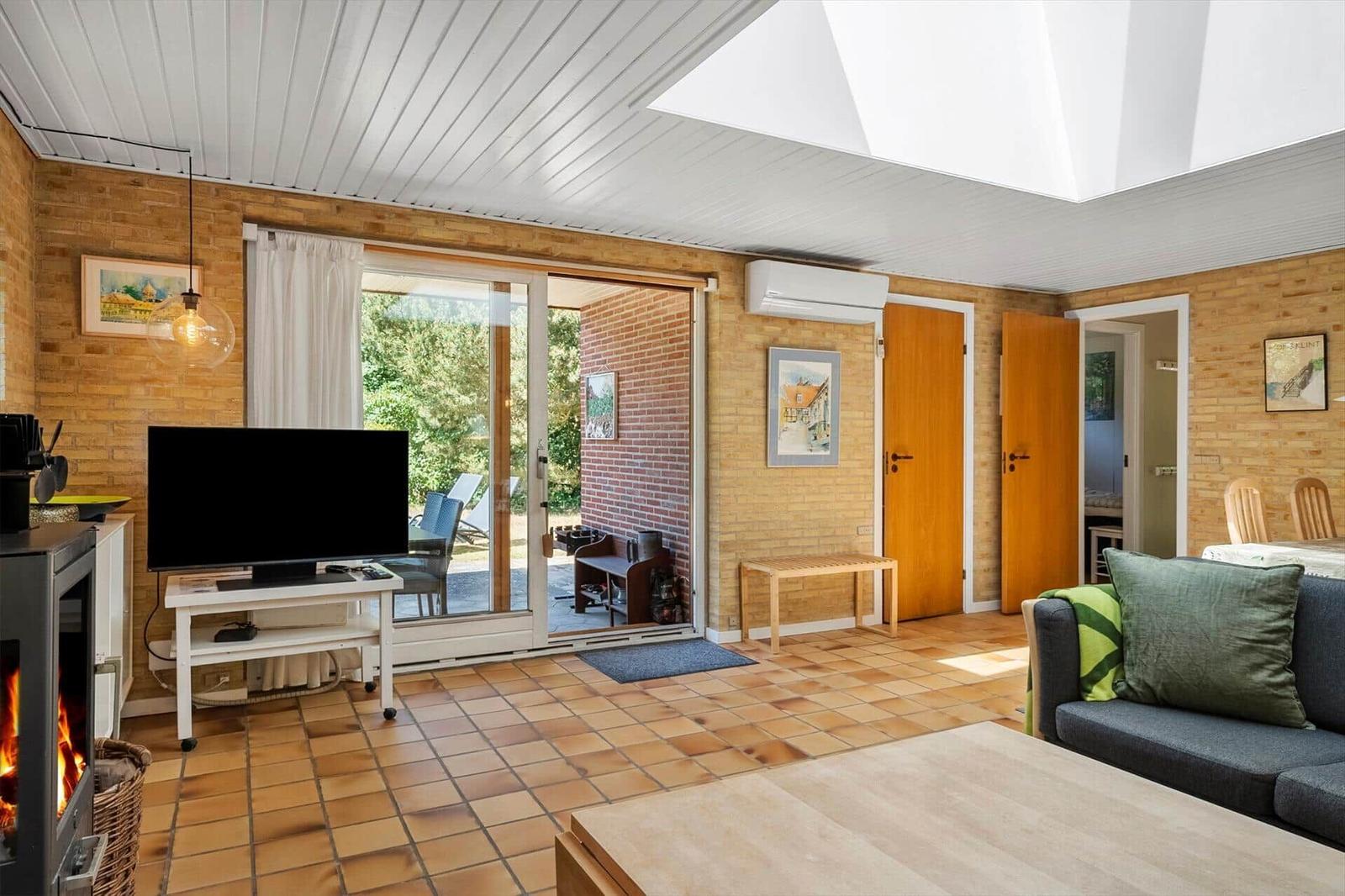 Wohnzimmer mit Kamin, TV und Terrasse. Holzwände und Terrakottaboden.