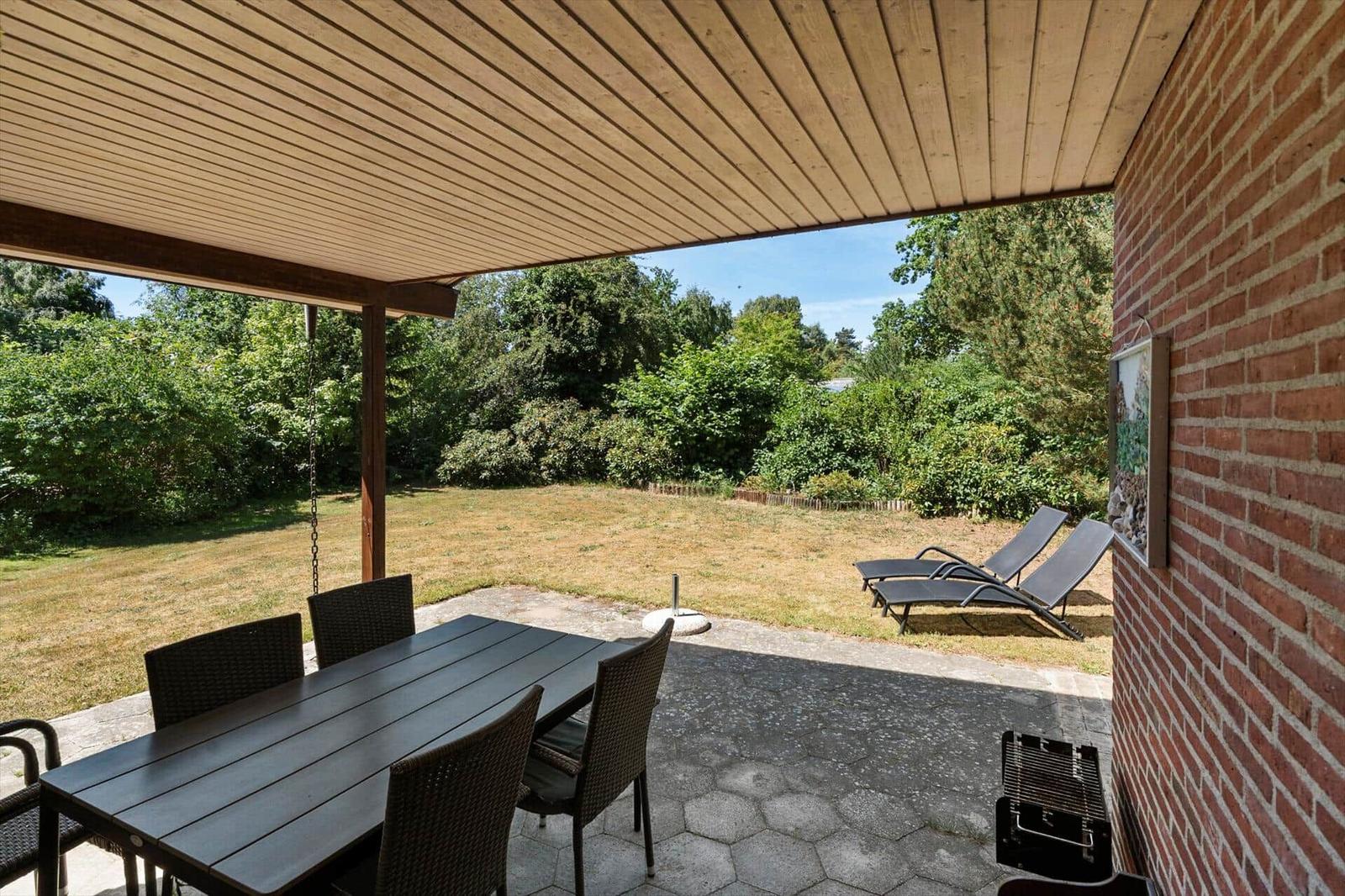 Terrasse mit Tisch, Stühlen und Liegen. Blick auf grünen Garten und Himmel.