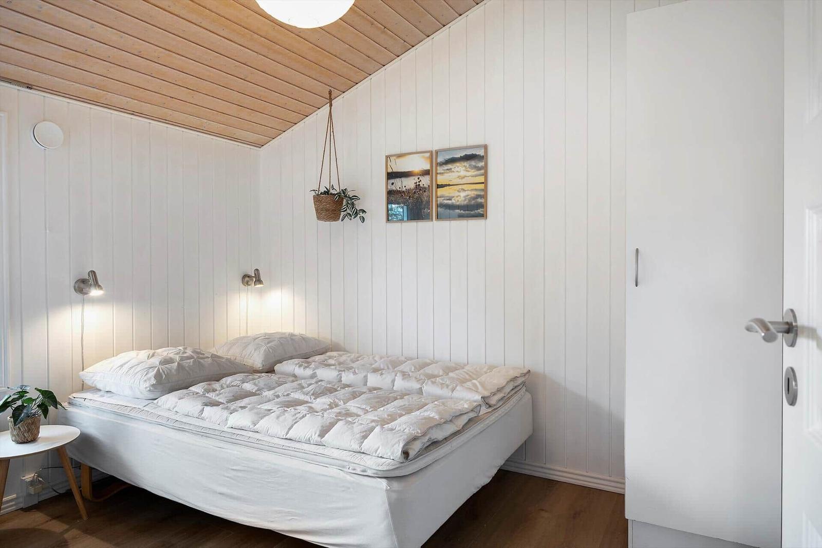 Schlafzimmer mit weißem Bett, Holzdecke und Wandpaneelen. Hängende Pflanze und Bilder an der Wand.