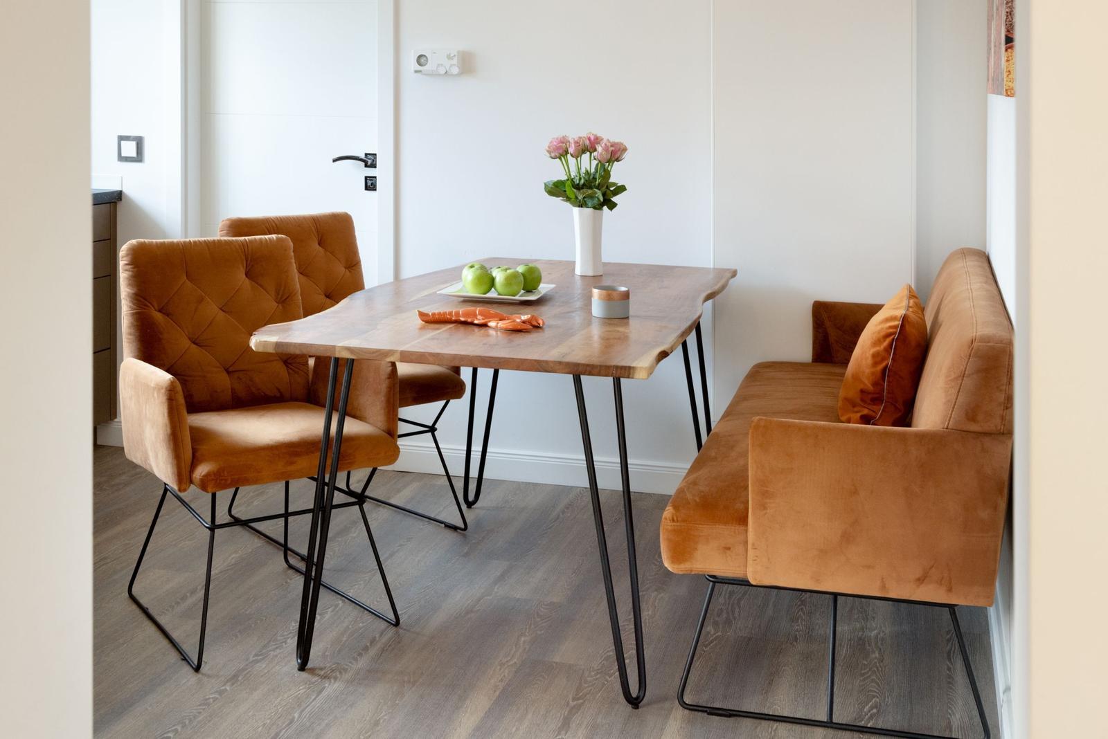 Esstisch mit zwei Stühlen und Sofa, Holzplatte, schwarze Beine, Blumen und Äpfel darauf.