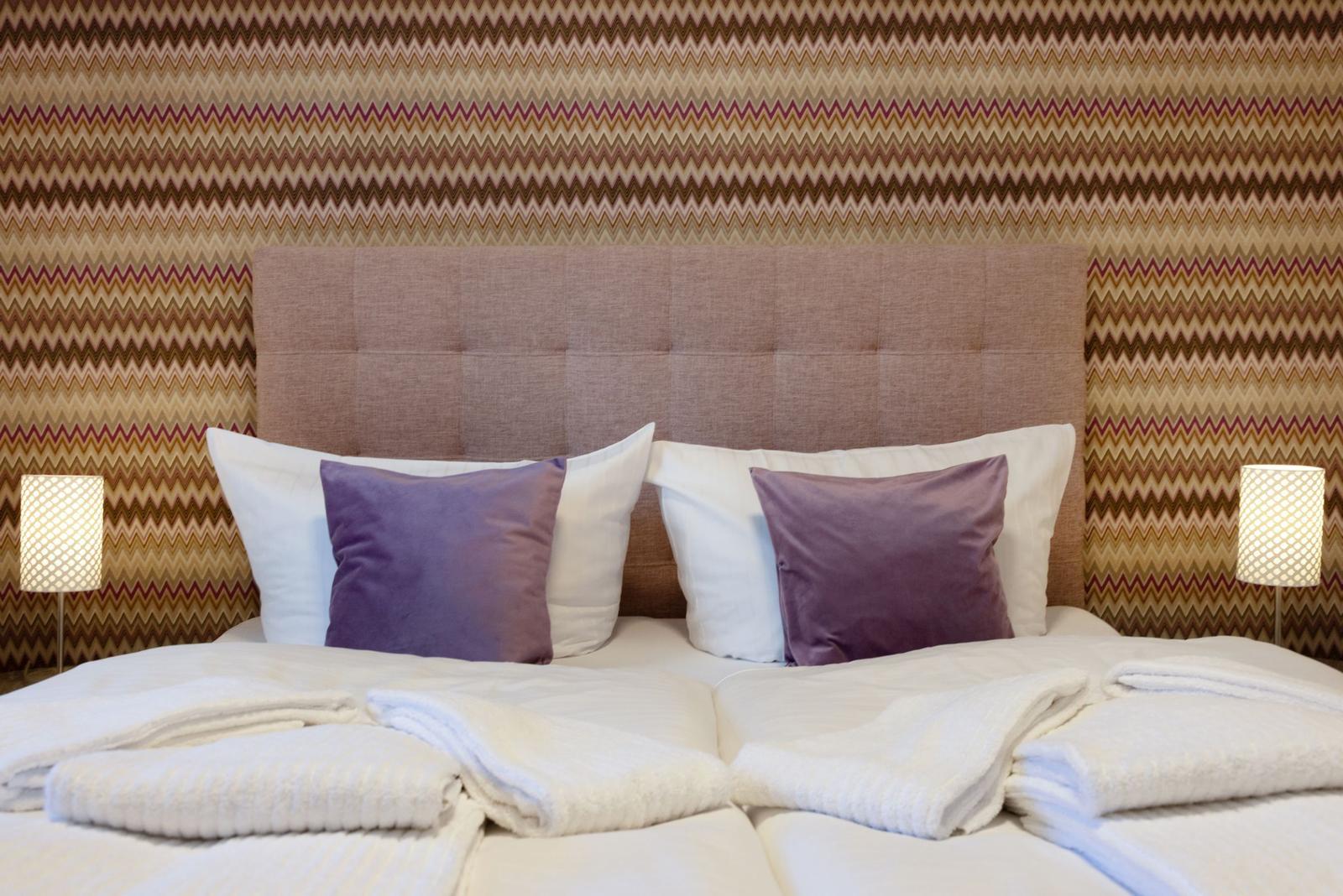 Ein Bett mit weißen Laken, zwei lila Kissen und zwei Nachttischlampen vor einem gemusterten Wandbezug.