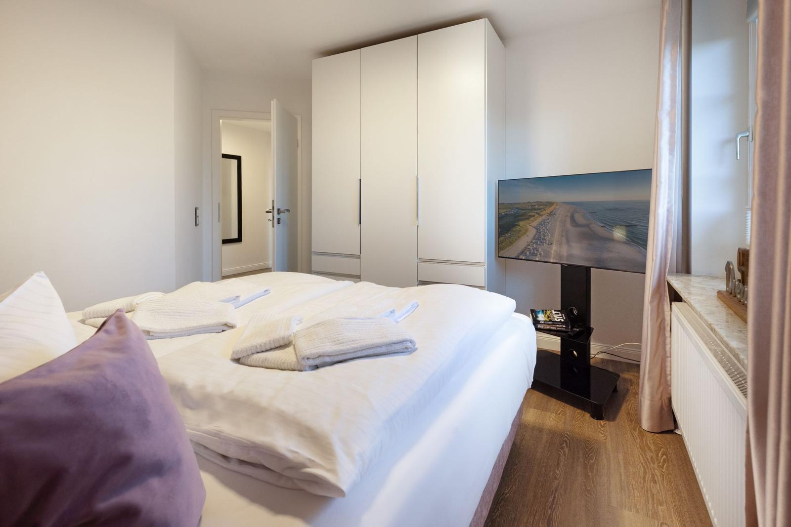 Schlafzimmer mit Bett, Kleiderschrank und Fernseher. Holzboden und Fenster mit Vorhängen.