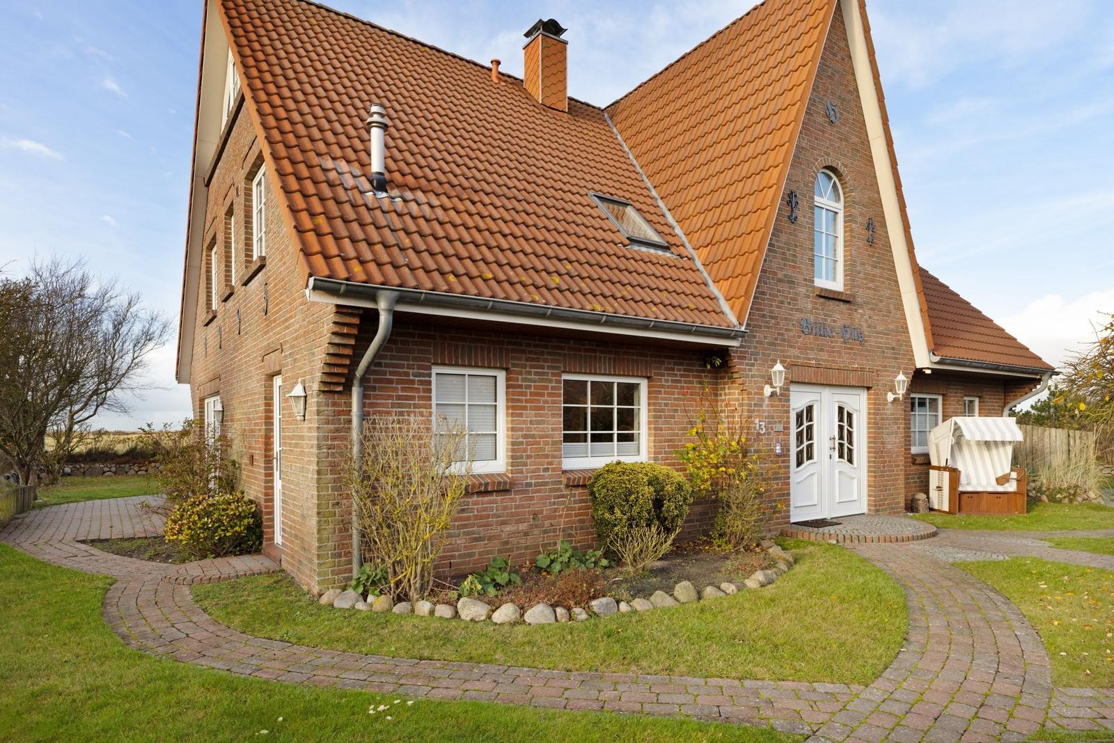 Ein zweistöckiges Haus mit rotem Ziegeldach und weißer Haustür. Vor dem Haus führt ein gepflasterter Weg.