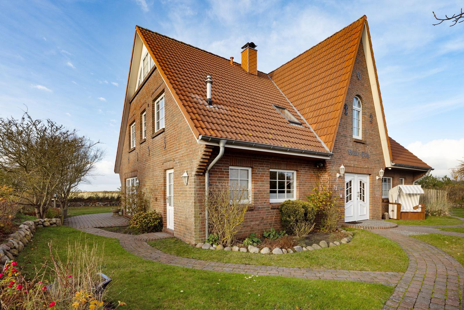 Ein zweistöckiges Haus mit rotem Ziegeldach und weißen Fenstern. Vorhof mit gepflastertem Weg und Grünfläche.