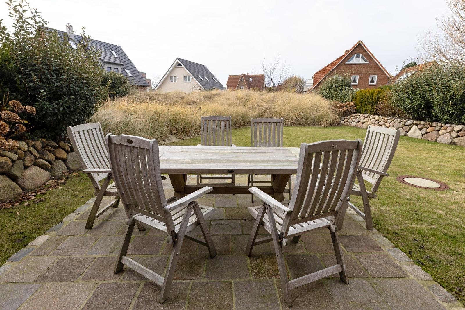 Holztisch mit sechs Stühlen auf Steinplatten im Garten. Hintergrund: Gras und Häuser.