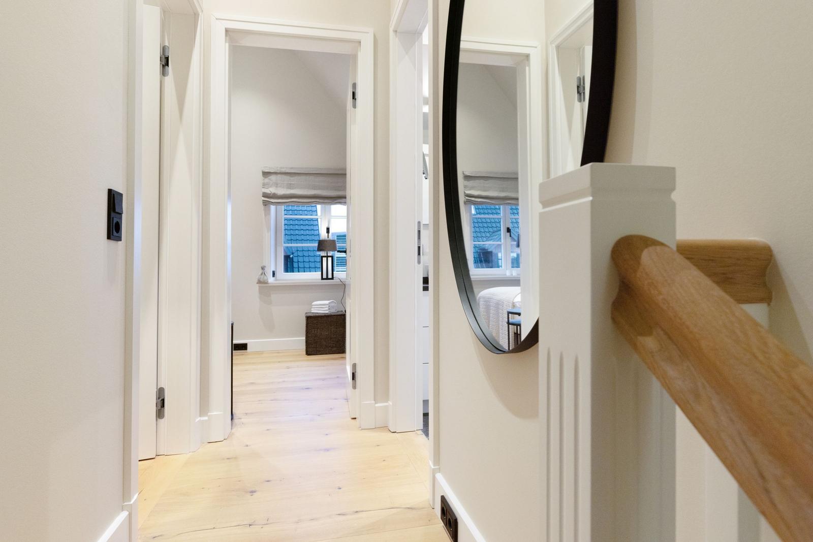 Flur mit Holzboden, Spiegel und Treppengeländer. Durchgang zu Schlafzimmer mit Fenster.