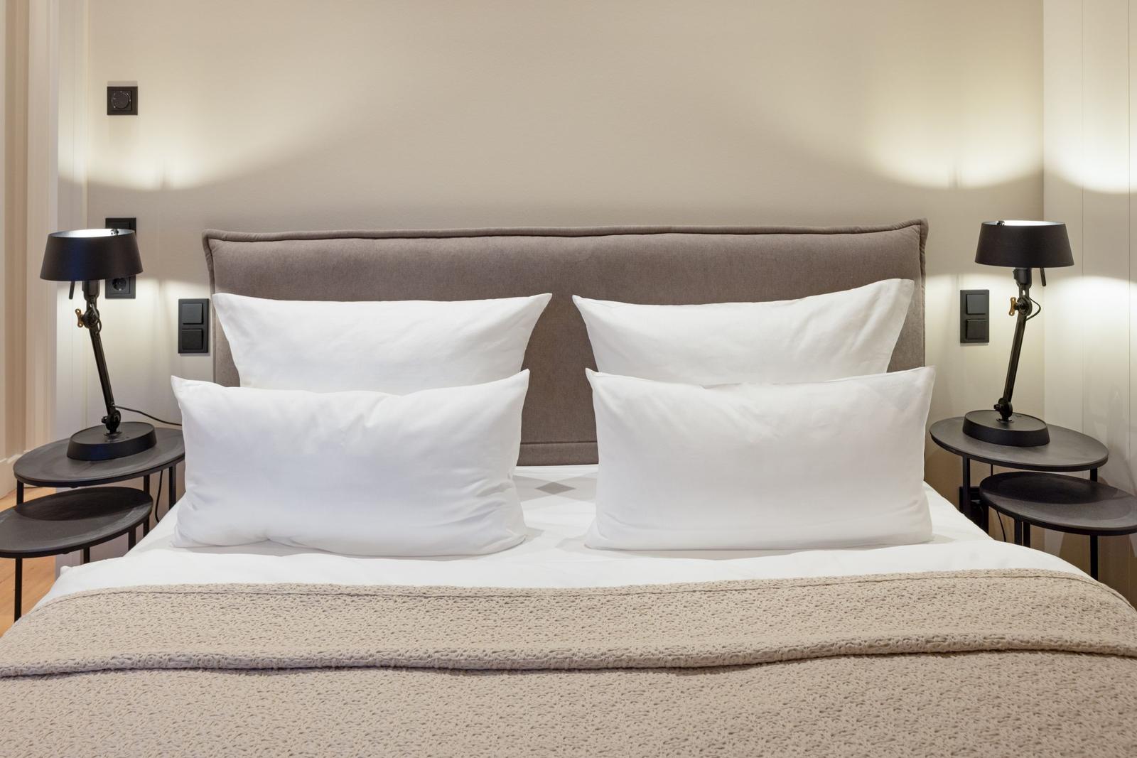 Ein Bett mit vier weißen Kissen und einer hellen Bettdecke. Zwei Nachttische mit schwarzen Lampen.