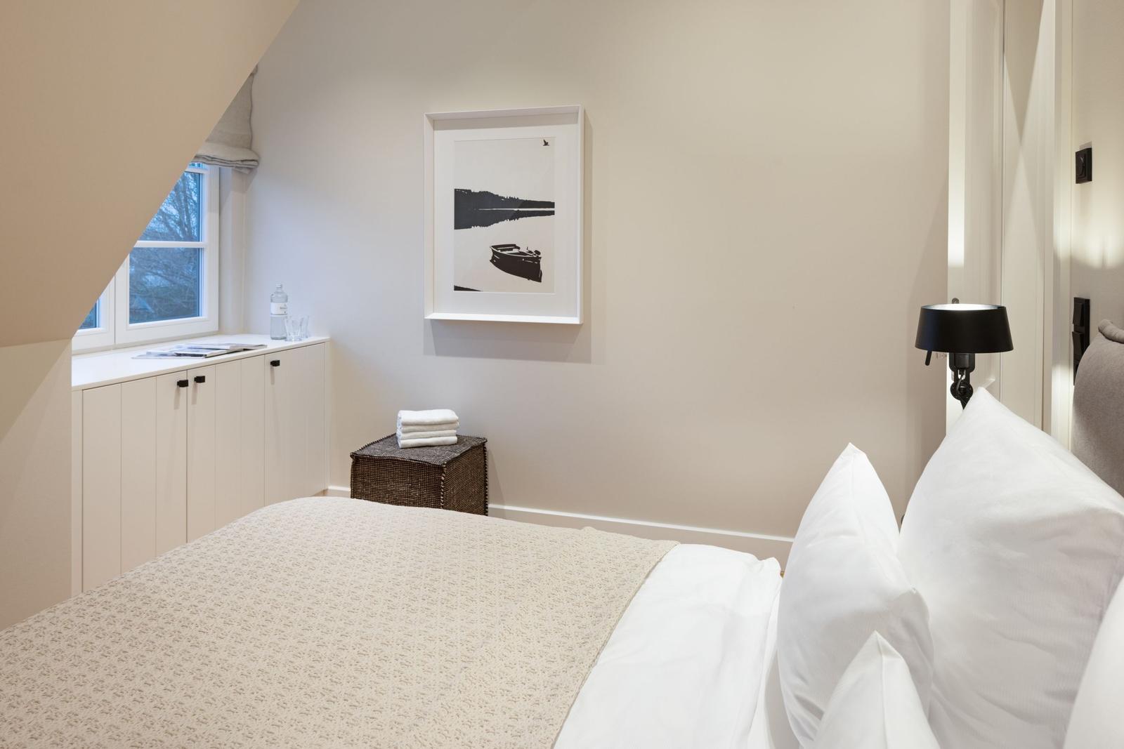 Schlafzimmer mit Bett, Nachttisch und Fenster. Wandbild und Wasserflasche sind sichtbar.
