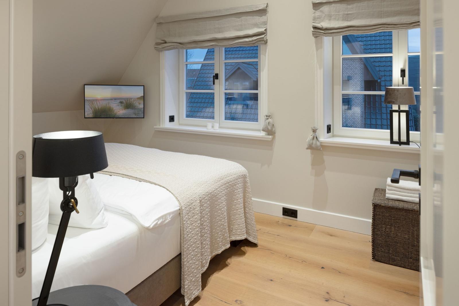 Schlafzimmer mit Bett, Fenstern und TV. Holzboden und neutral eingerichtet.