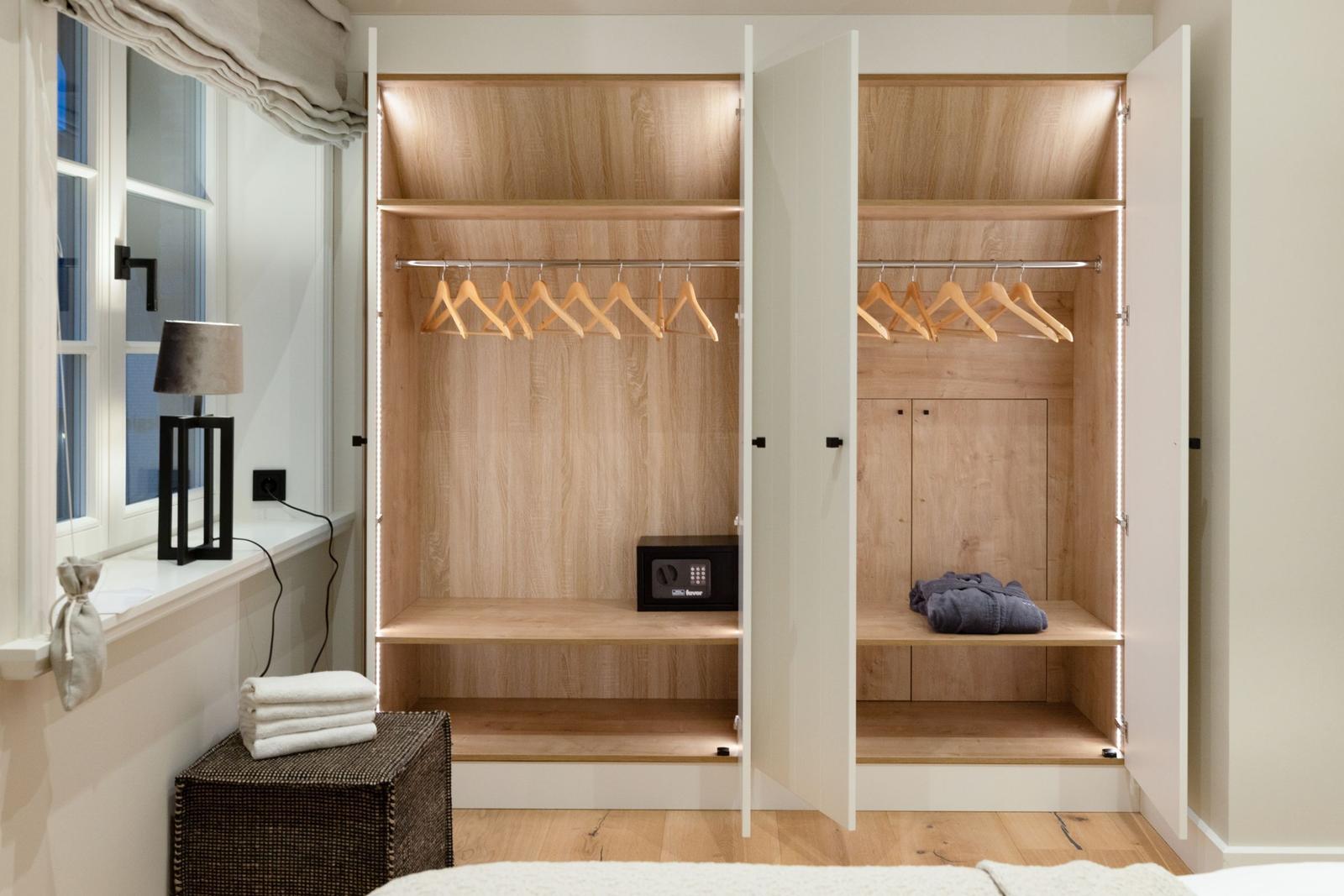 Schrank mit Holzregalen, Haken und Safe. Fenster, Lampe und Handtuchkorb sichtbar.
