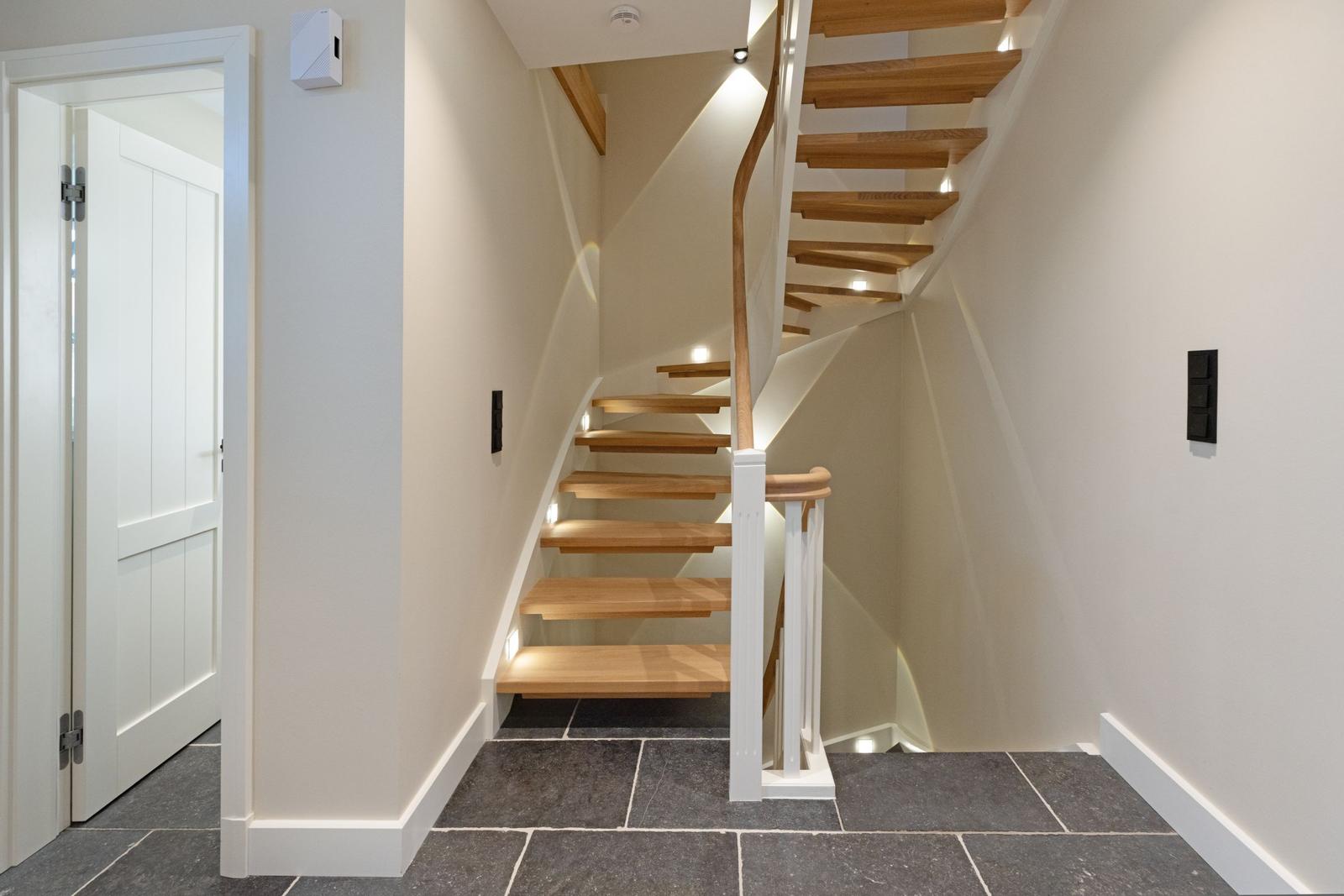 Holzgetreppe mit weißem Geländer führt nach oben. Dunkle Fliesenboden und weiße Wände.