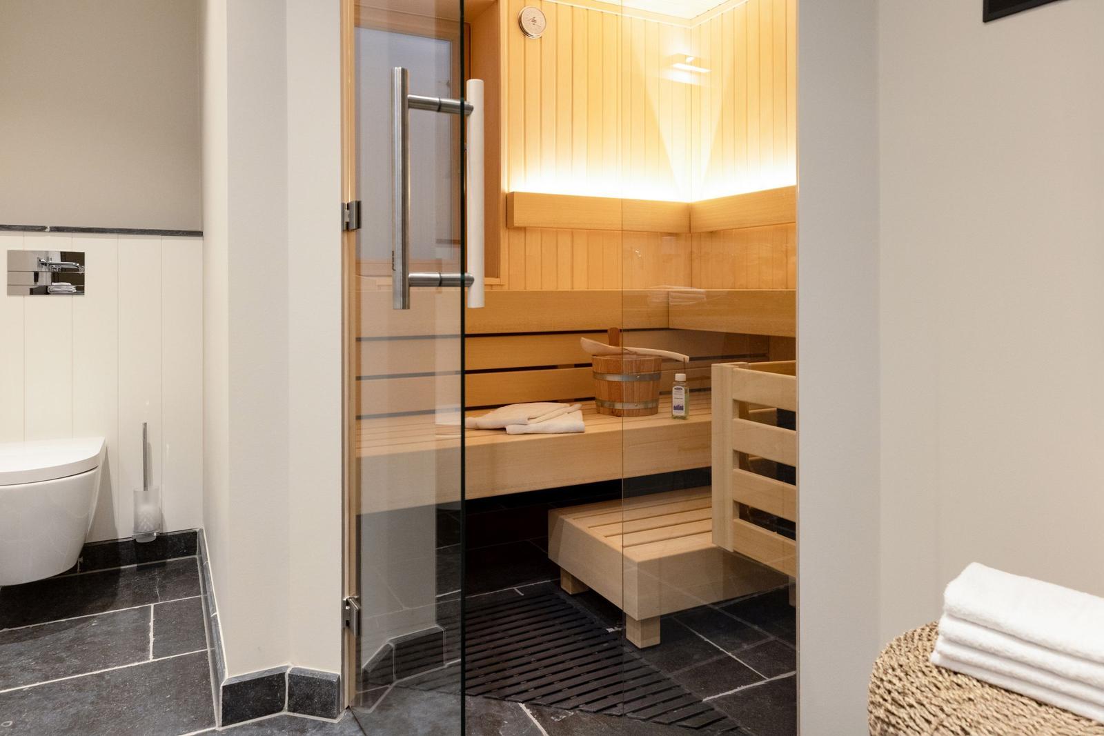 Sauna mit Holzbänken und Glaswand. Weiße Handtücher und Holzwaschbecken sind sichtbar.