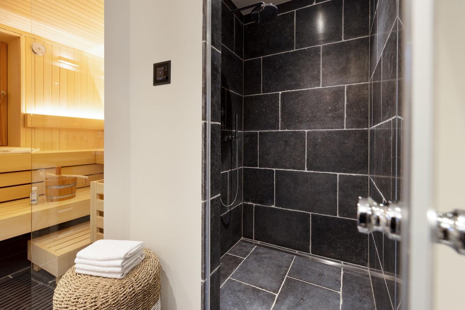Dusche mit schwarzen Fliesen und Sauna mit Holzbank im Hintergrund.