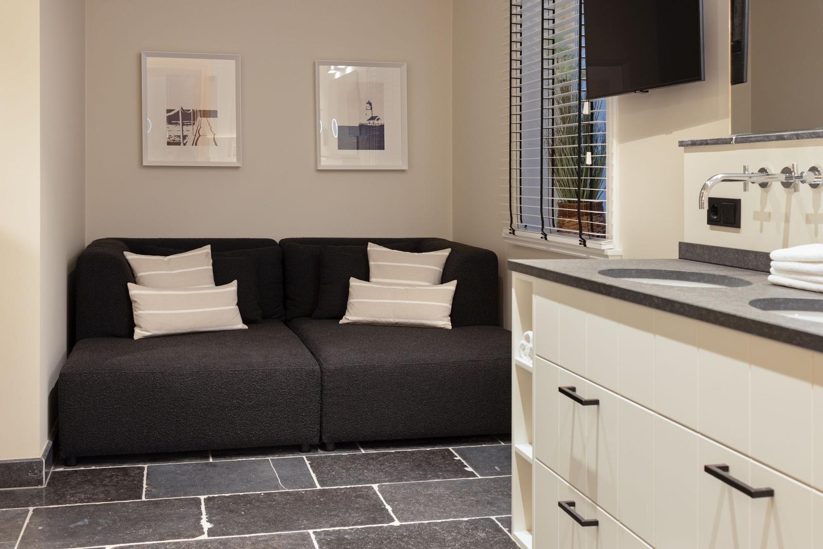 Schwarzes Sofa mit Kissen, Wandbilder, Fenster mit Jalousie und Waschtisch mit Spiegel.