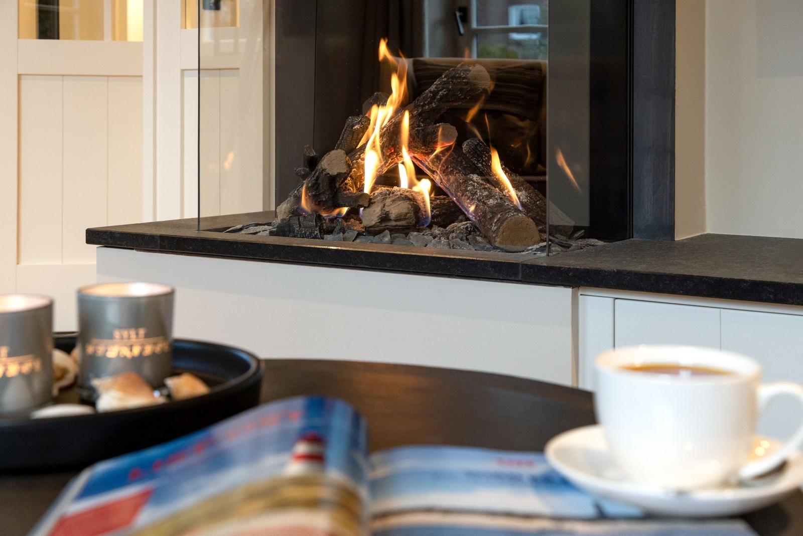 Ein Kamin mit brennenden Holzscheiten, eine Tasse Kaffee und ein geöffnetes Magazin auf einem Tisch.