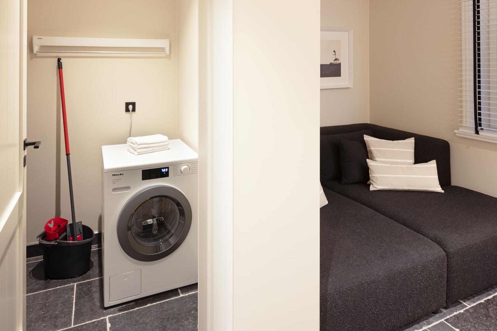 Wäscherei mit Waschmaschine und Reinigungsmitteln. Nebenraum mit Sofa und Fenster.