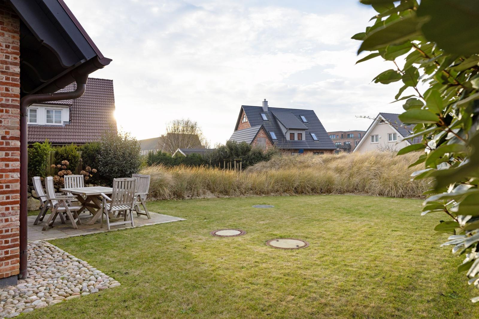 Hinterhof mit Garten, Tisch und Stühlen. Im Hintergrund sind Häuser und Grasland zu sehen.
