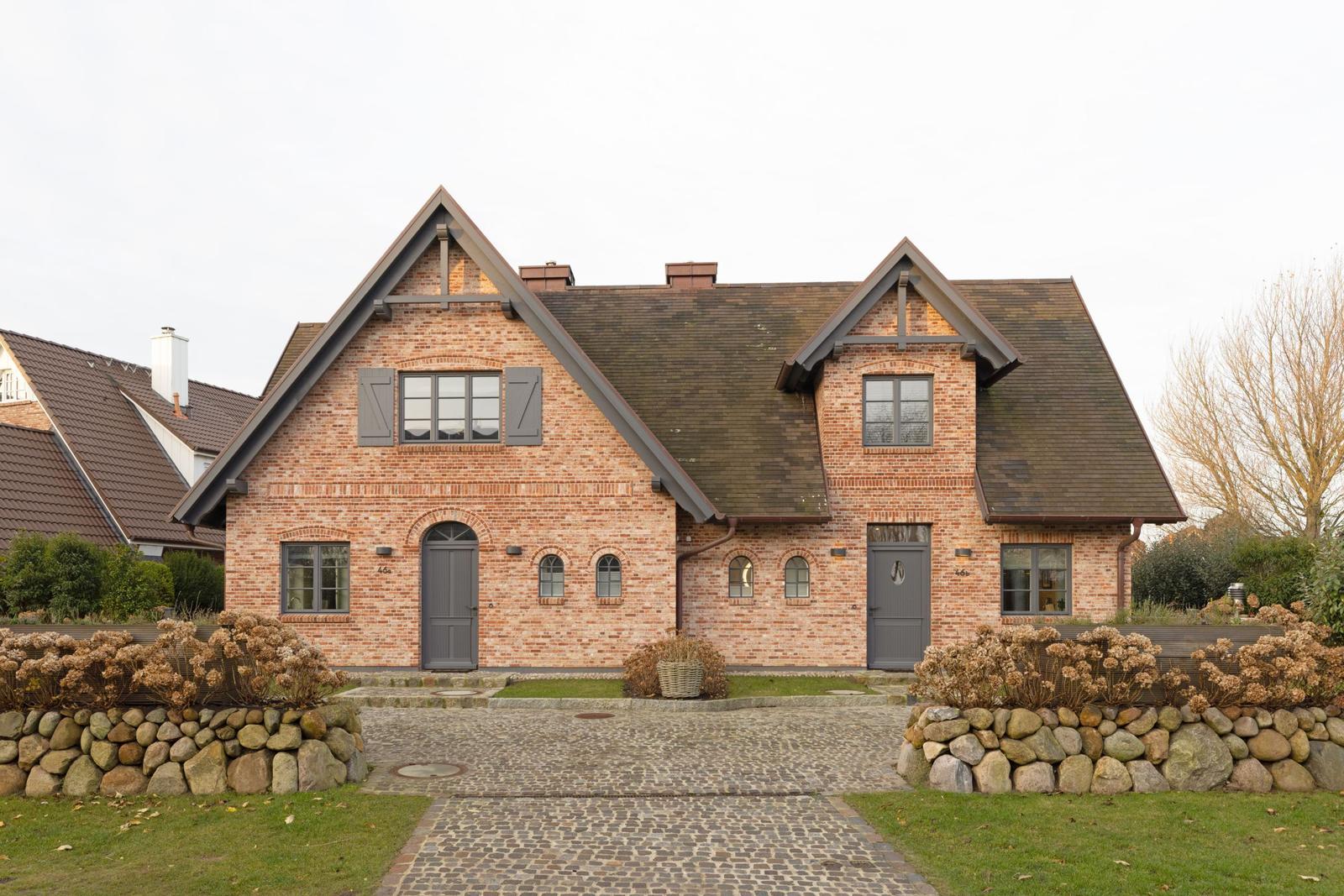 Haus mit Backsteinfassade, grauen Fenstern und Dachgauben. Kiesweg und Steinmauer im Vorgarten.