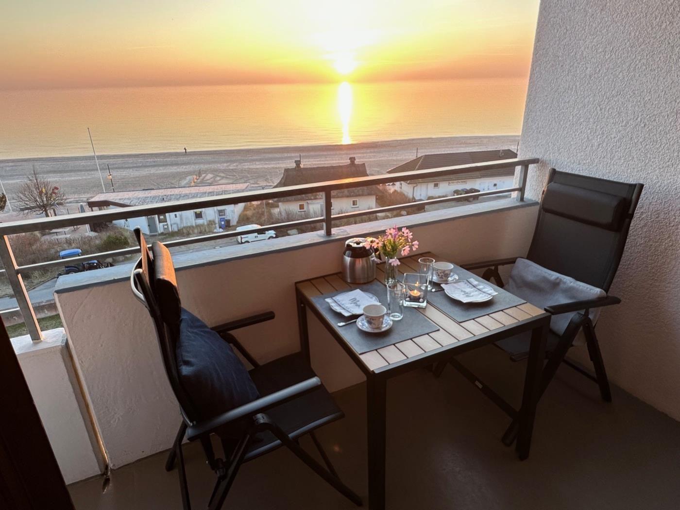 Balcony with table and chairs overlooking sunset over sea and beach.