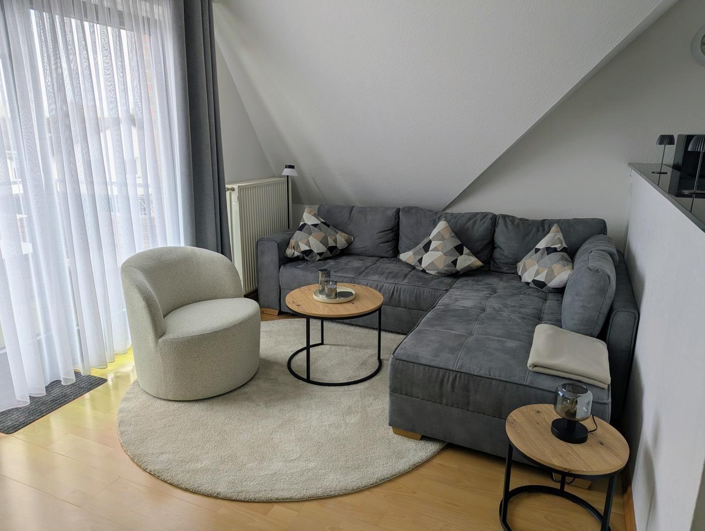 Wohnzimmer mit grauem Sofa, runden Tischen und großem Fenster mit weißen Vorhängen.