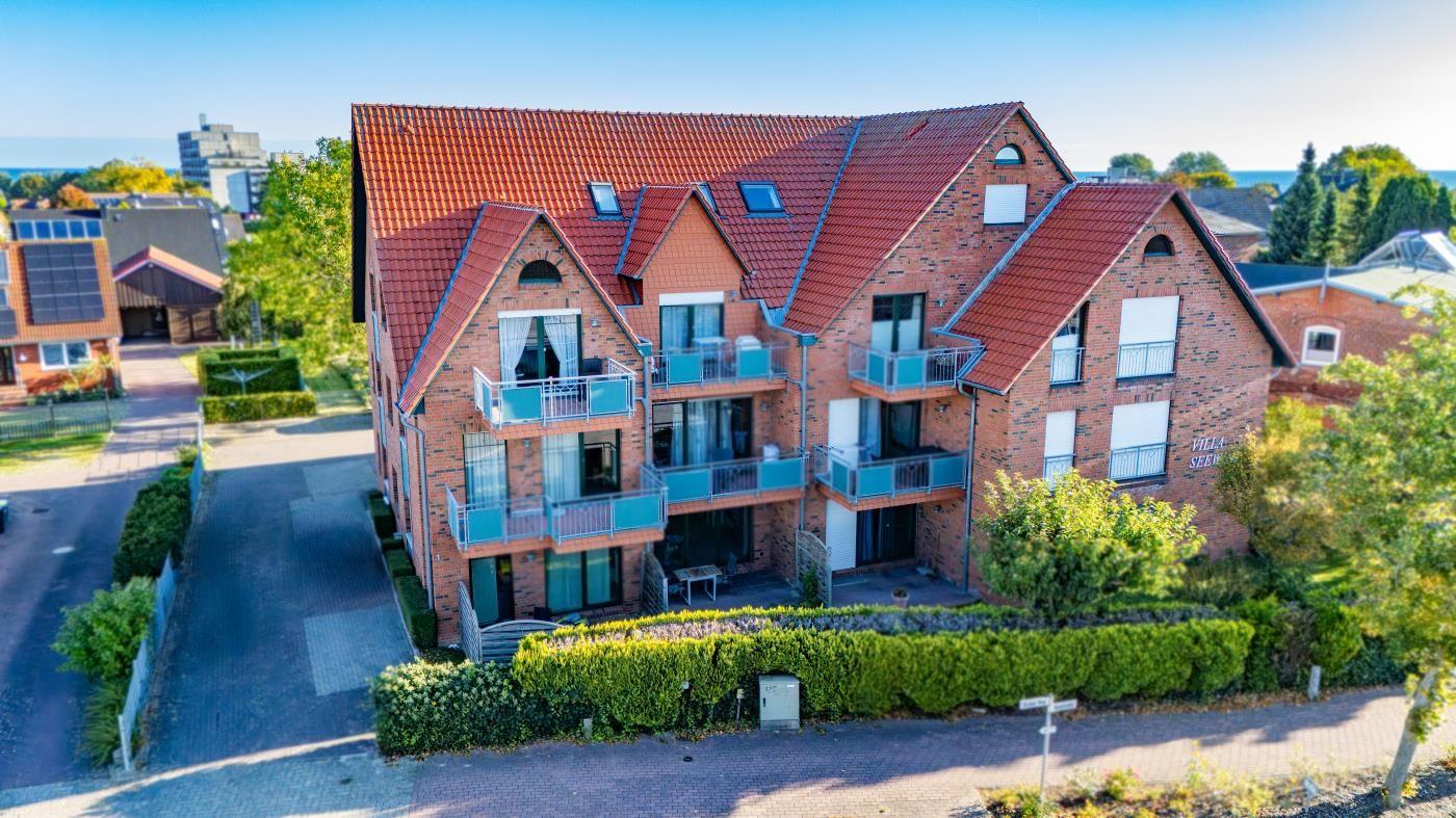 Zweistöckiges Backsteinhaus mit Balkonen und rotem Dach. Umgeben von Grünflächen und Bäumen.