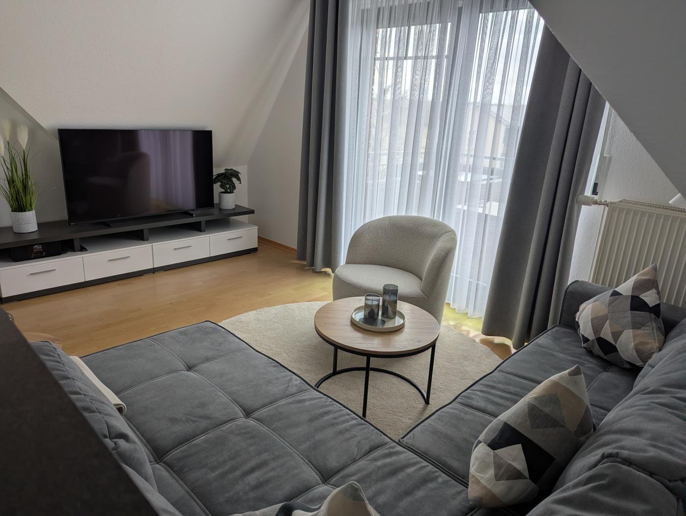 Wohnzimmer mit Sofa, Sessel, Fernseher und großem Fenster mit Vorhängen.