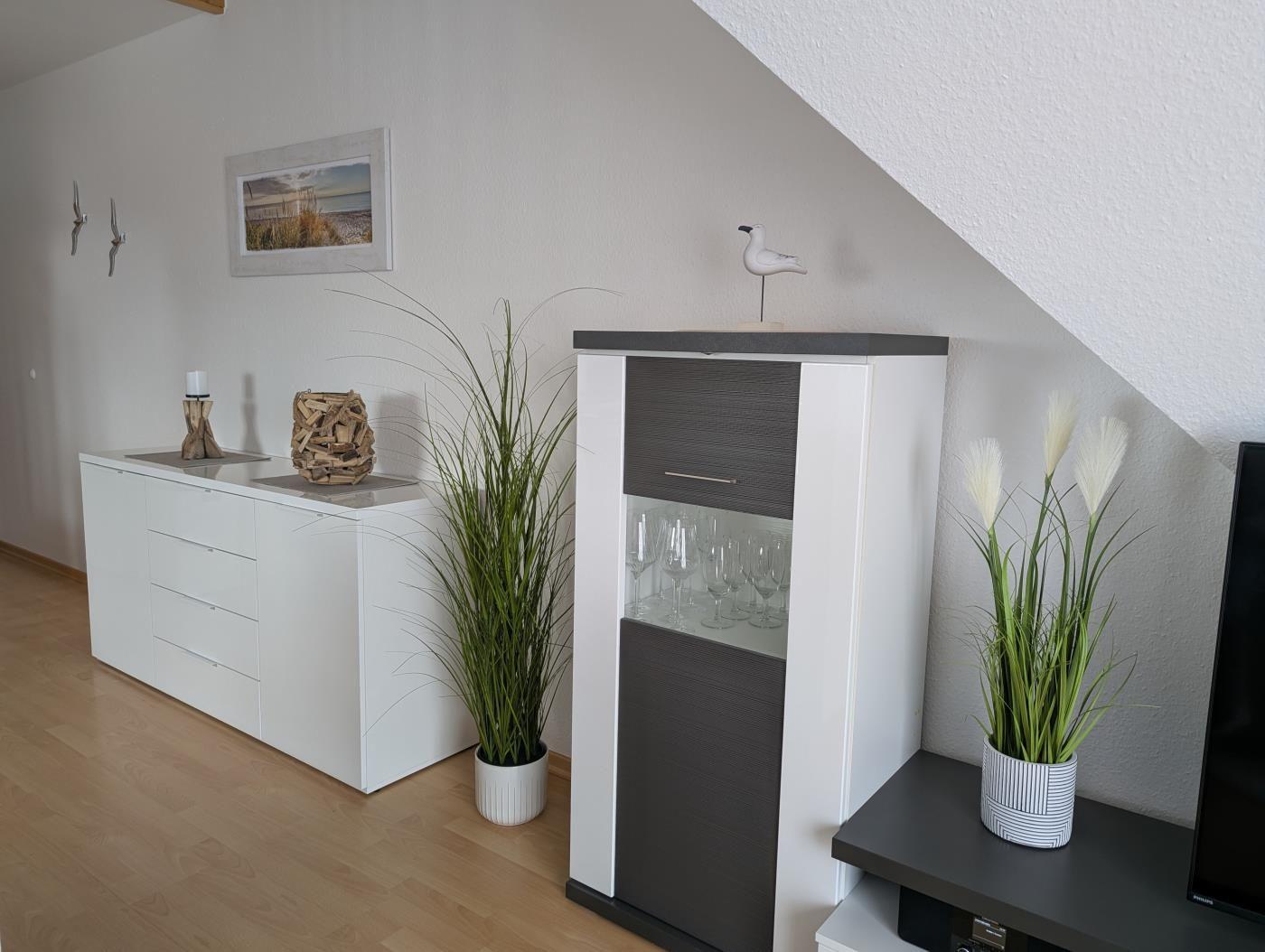 Wohnzimmer mit weißen Schränken, Pflanzen und Glasregal unter schräger Decke.