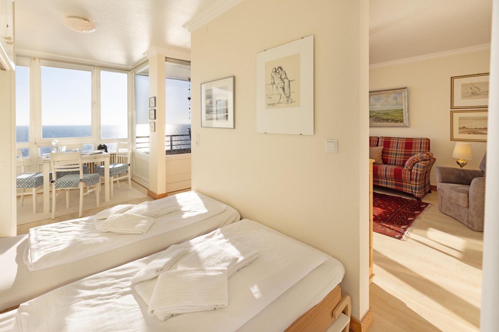 Zwei Betten im Schlafzimmer. Durchgang in Wohnzimmer mit Sofa und Blick aufs Meer.