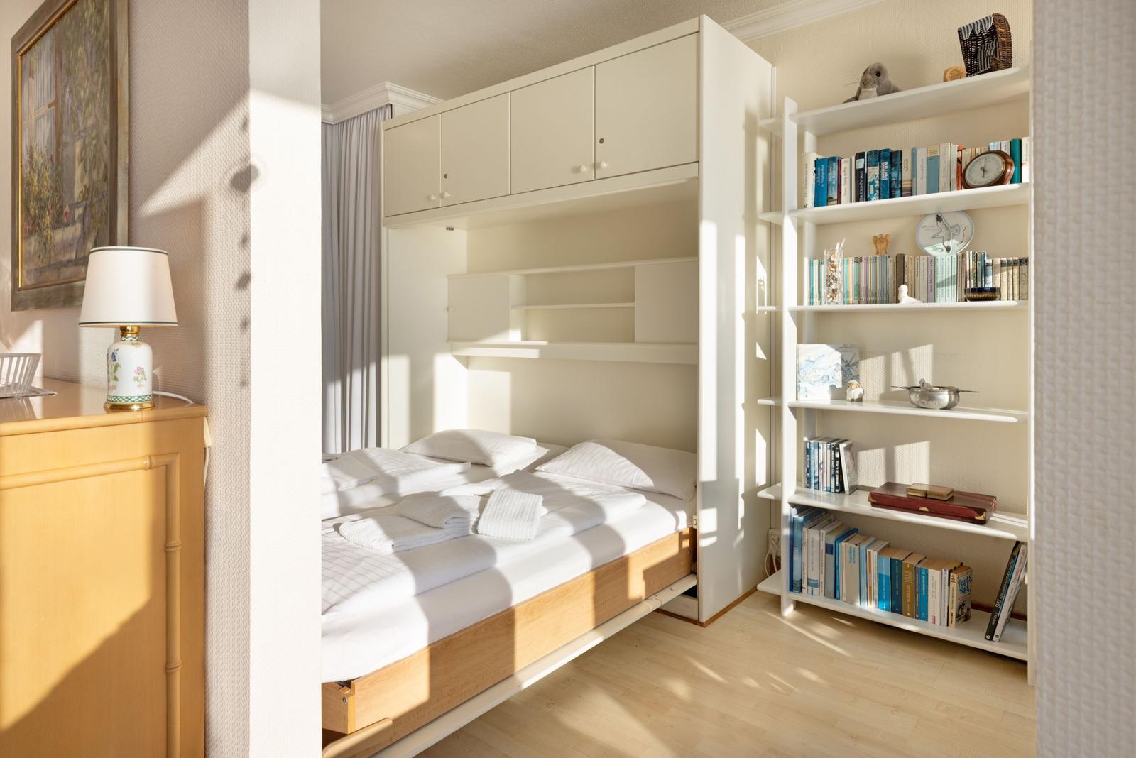 Schlafzimmer mit Bett, Bücherregal und Schrank. Sonnenlicht fällt durch Fenster.