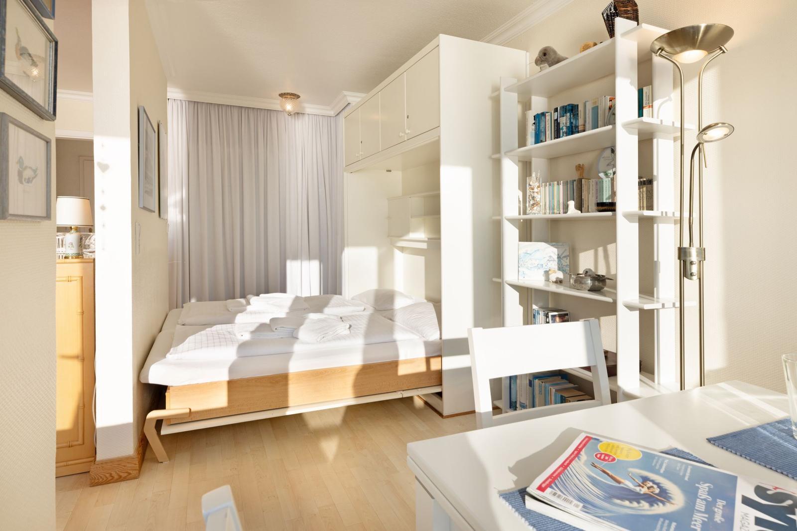 Schlafzimmer mit Bett, Bücherregal und Tisch. Fenster mit Vorhängen. Holzboden und weiße Möbel.