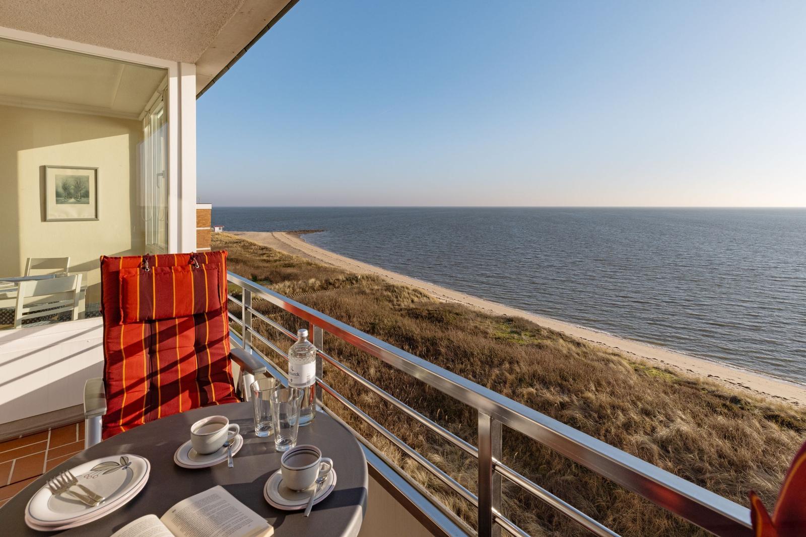 Balkon mit Tisch, Stühlen und Meerblick. Roter Sessel, Tisch mit Tassen und Flasche.