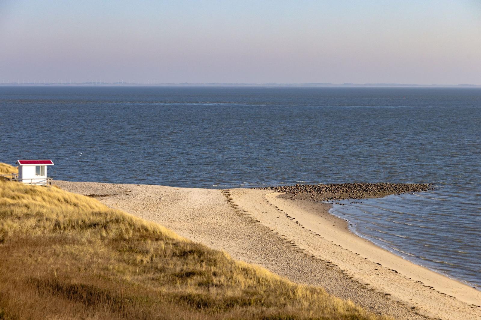 Strand mit Sand, Gras und kleinem Haus am Wasser.